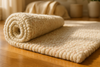 A premium rolled wool rug partially unrolled on a clean wooden floor — close-up of thick, plush wool pile with visible density and fine fibers, a soft beige and ivory color palette with light pink accents, natural light streaming in from a window, styled with cozy throws and neutral décor — sophisticated and high-end look.
