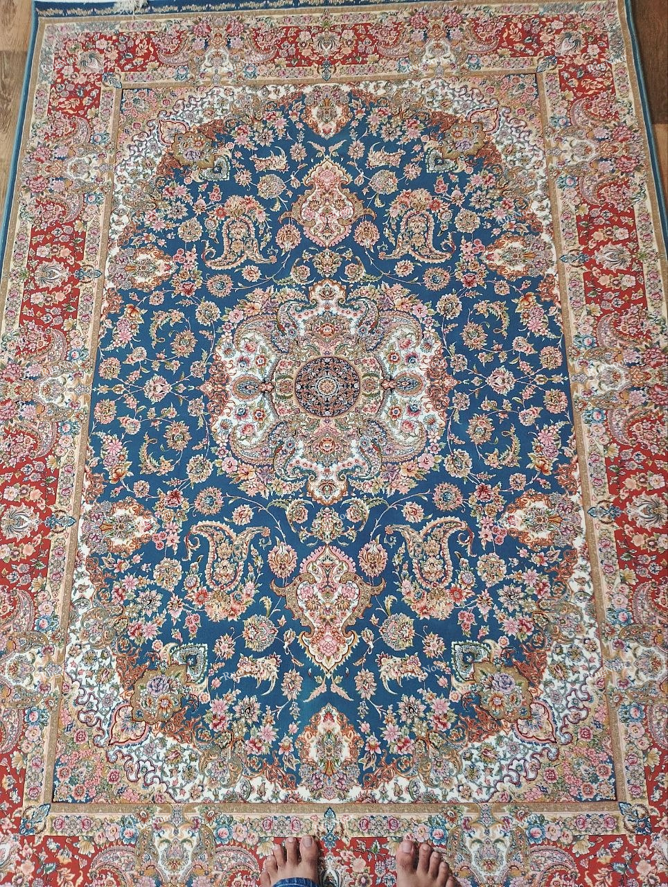 RugNoor Ruvaira 5x7 traditional floral carpet with a teal-blue centre, ornate floral medallion, rust-red border, ivory and blush-pink details, and decorative fringe.