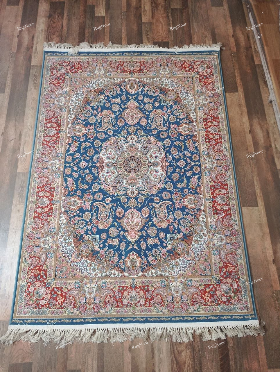 RugNoor Ruvaira 5x7 traditional floral carpet with a teal-blue centre, ornate floral medallion, rust-red border, ivory and blush-pink details, and decorative fringe.