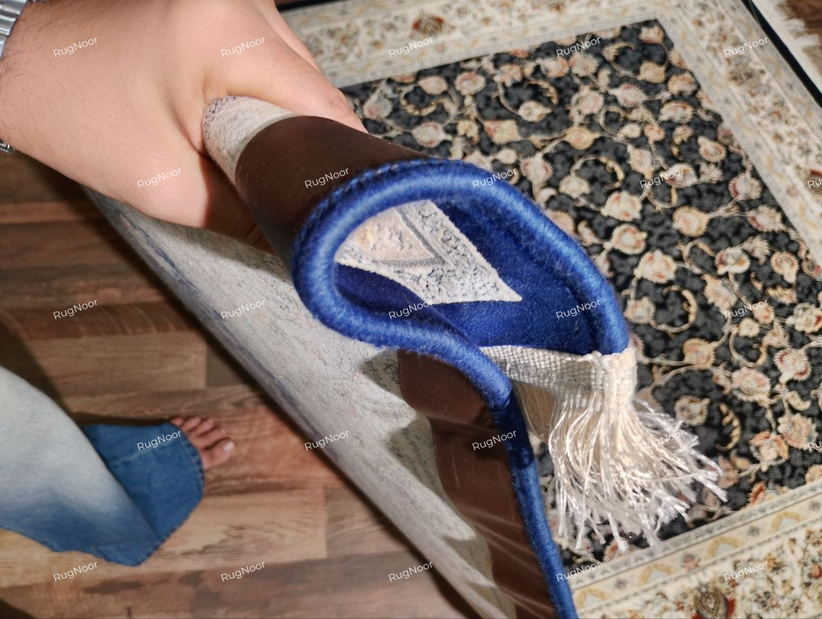 Folded corner of the Zarniva traditional carpet showing its brown underside, blue bound edge, ivory fringe and navy blue floral pattern.