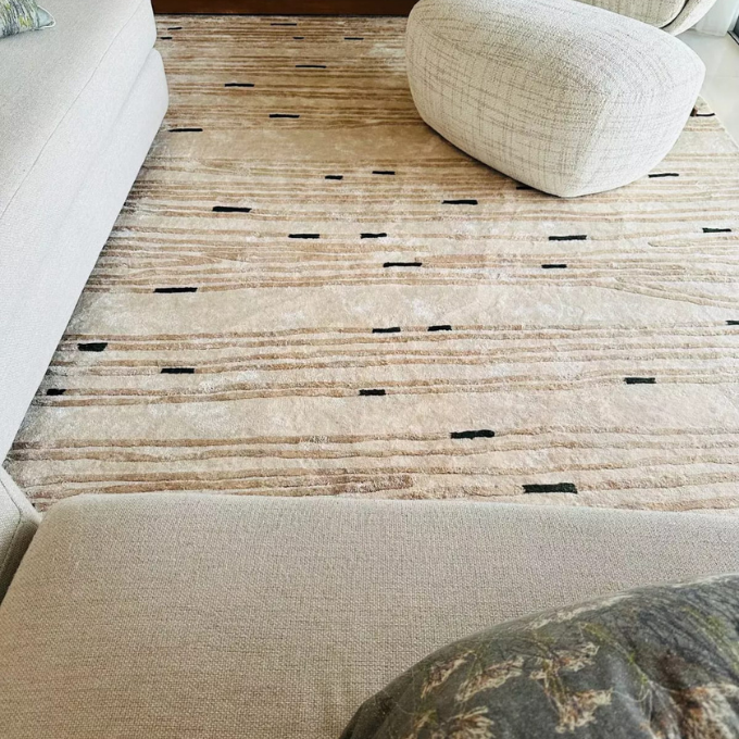 A beige and cream textured area rug with horizontal woven stripes and small black dash patterns, styled in a living room setting with a white sofa, a cream pouf, and a patterned throw pillow.