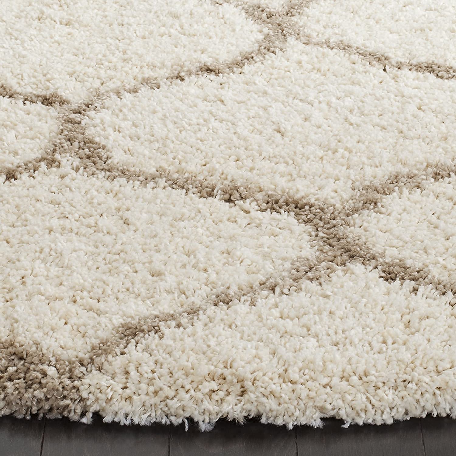 Close-up of a shag area rug with a cream and beige geometric trellis pattern, showing the plush texture and the edge of the rug resting on a dark floor.