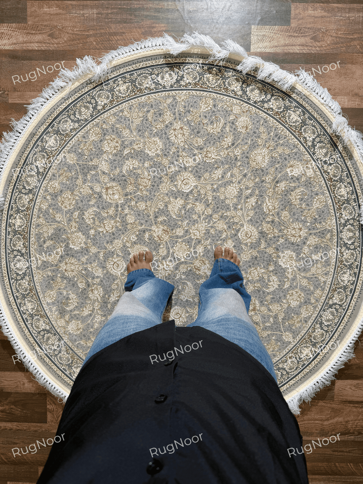 Golnaré round carpet shown with a person standing on it, displaying the grey floral field, delicate gold motifs, and dark ornamental border.