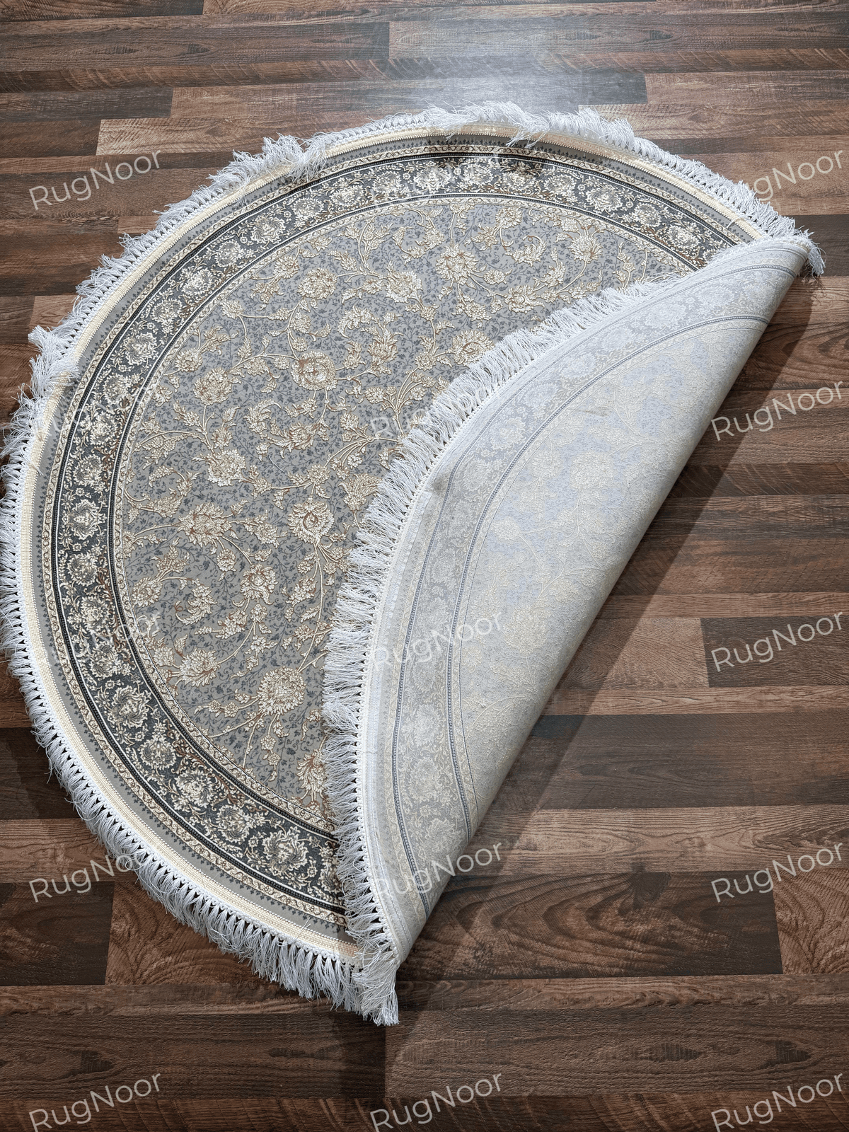 Golnaré grey round carpet with a corner folded back, revealing the pale underside alongside the floral pattern and ivory fringe.