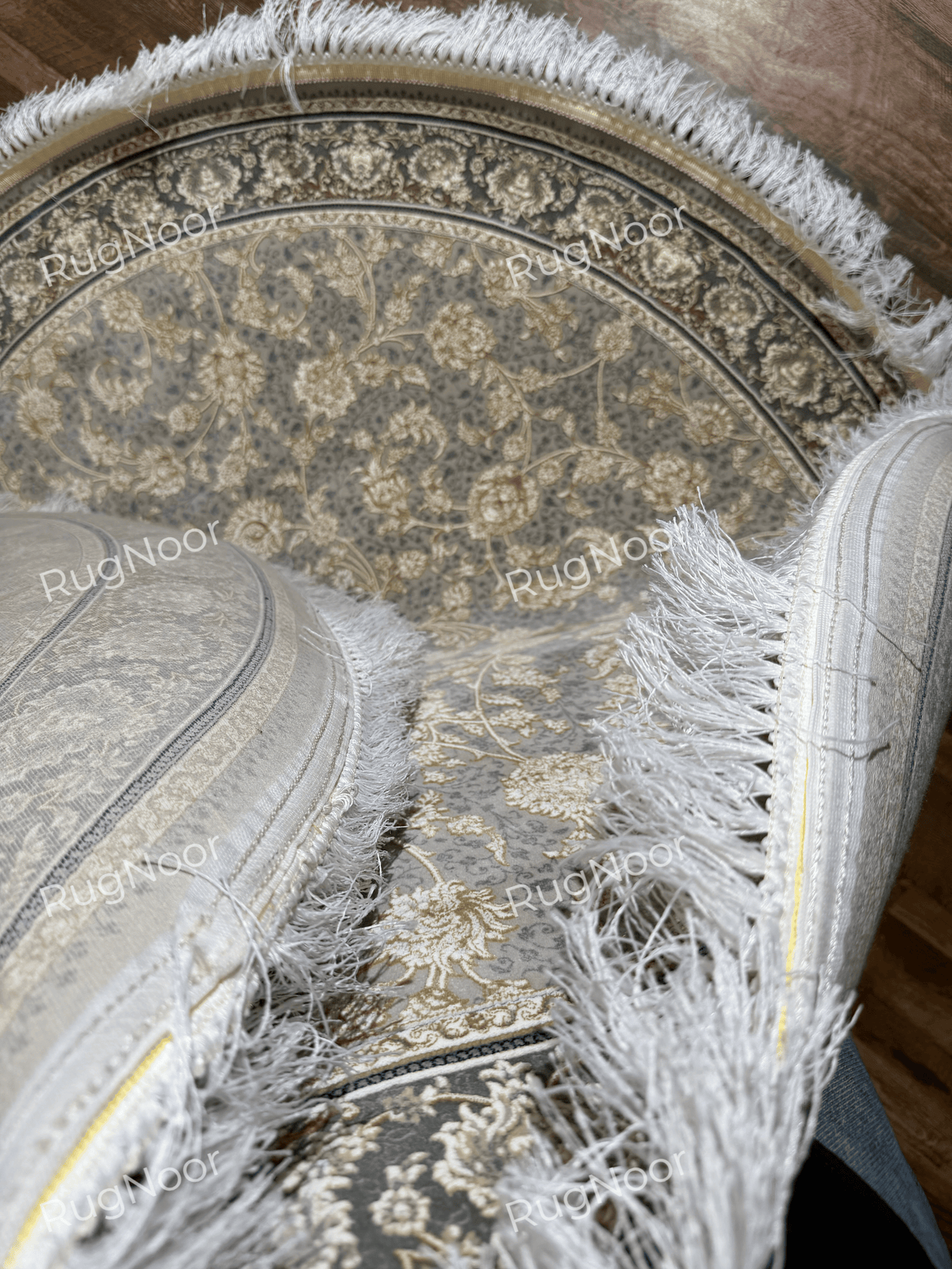 Golnaré carpet folded into a curved shape, showing its grey floral pattern, dark border, ivory fringe, and finished edge.