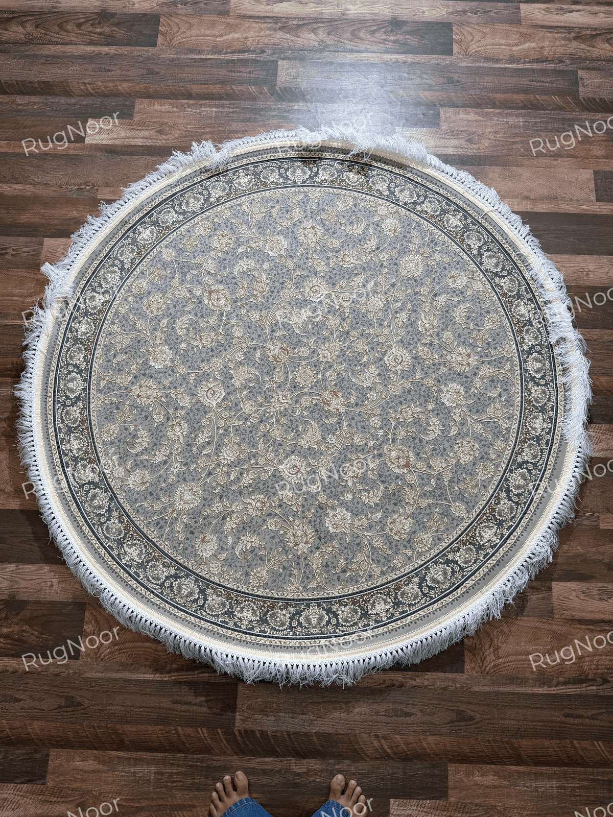 Full overhead view of the Golnaré round carpet with an all-over ivory and gold floral pattern on grey, framed by a dark ornamental border and ivory fringe.