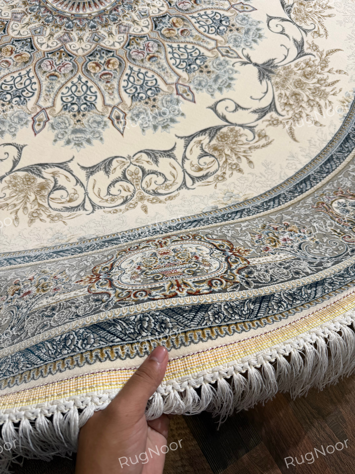 Mahzarin round carpet lifted into a curve to show the patterned surface, ivory fringe, and layered border along the edge.