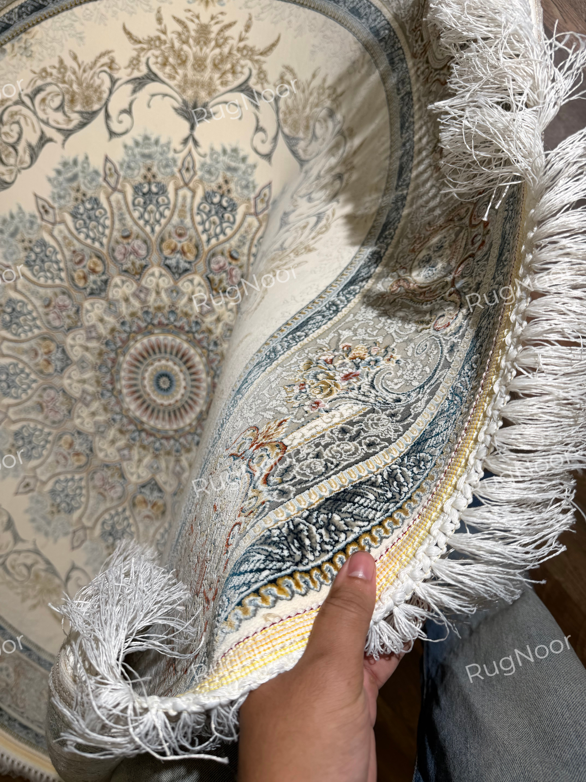 Mahzarin round carpet lifted into a curve to show the patterned surface, ivory fringe, and layered border along the edge.