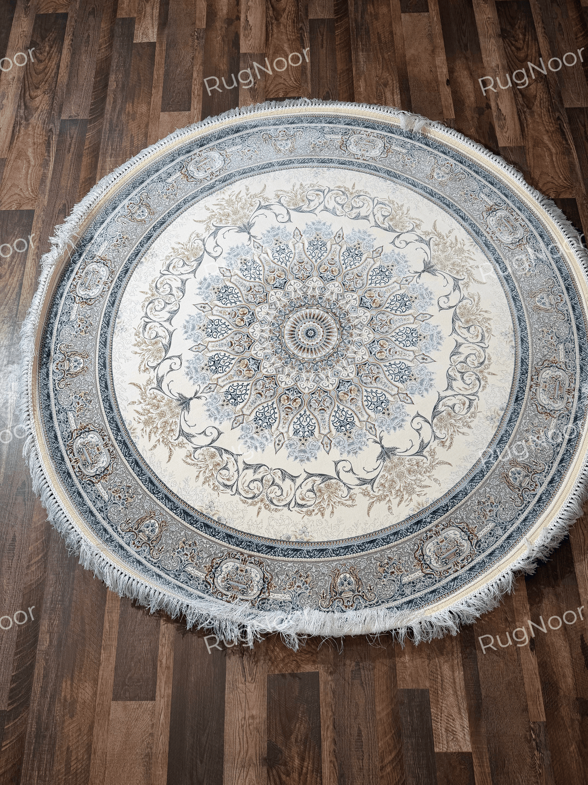Full view of the Mahzarin round ivory carpet with a detailed central medallion, blue-grey floral border, muted gold accents, and ivory fringe on a wooden floor.