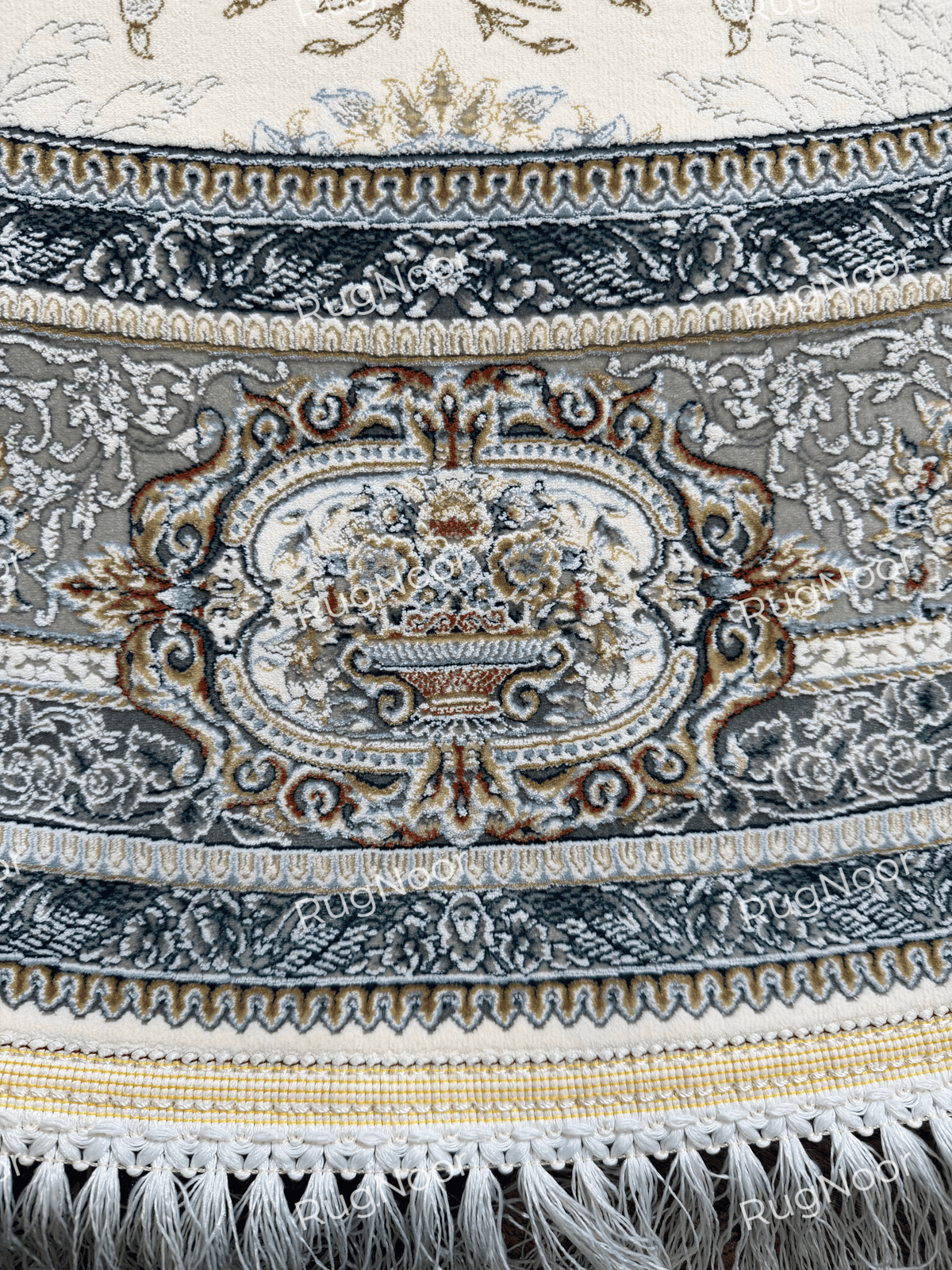 Close-up of the Mahzarin carpet’s grey ornamental border, small floral motifs, gold accents, woven edge, and ivory fringe.