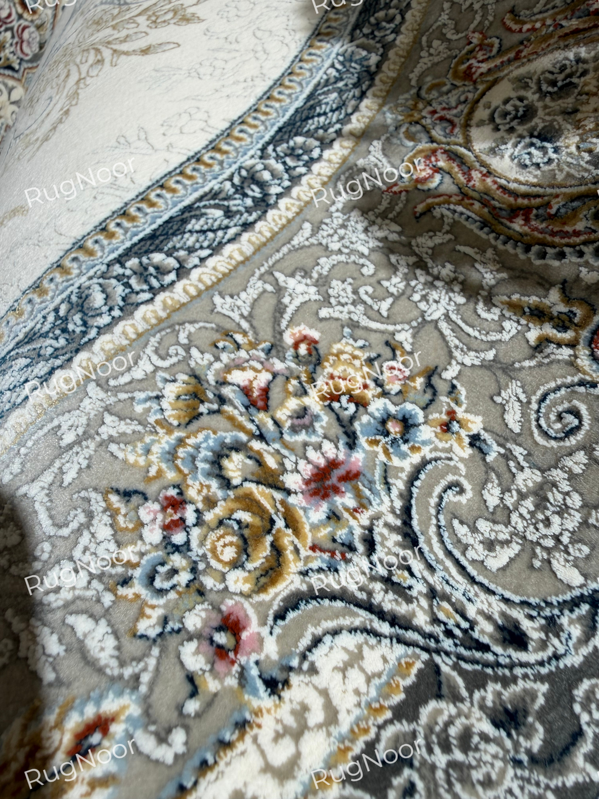 Close-up of taupe and grey floral scrollwork with cream, rust, and gold accents in the Mahzarin carpet’s ornamental border.