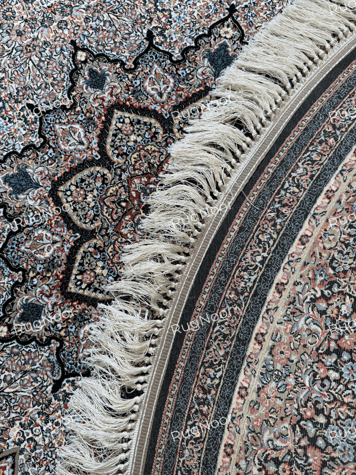 Close-up of the Ravzaran round carpet showing ivory fringe, a black ornamental band, and intricate rust, ivory, and blue floral detailing.