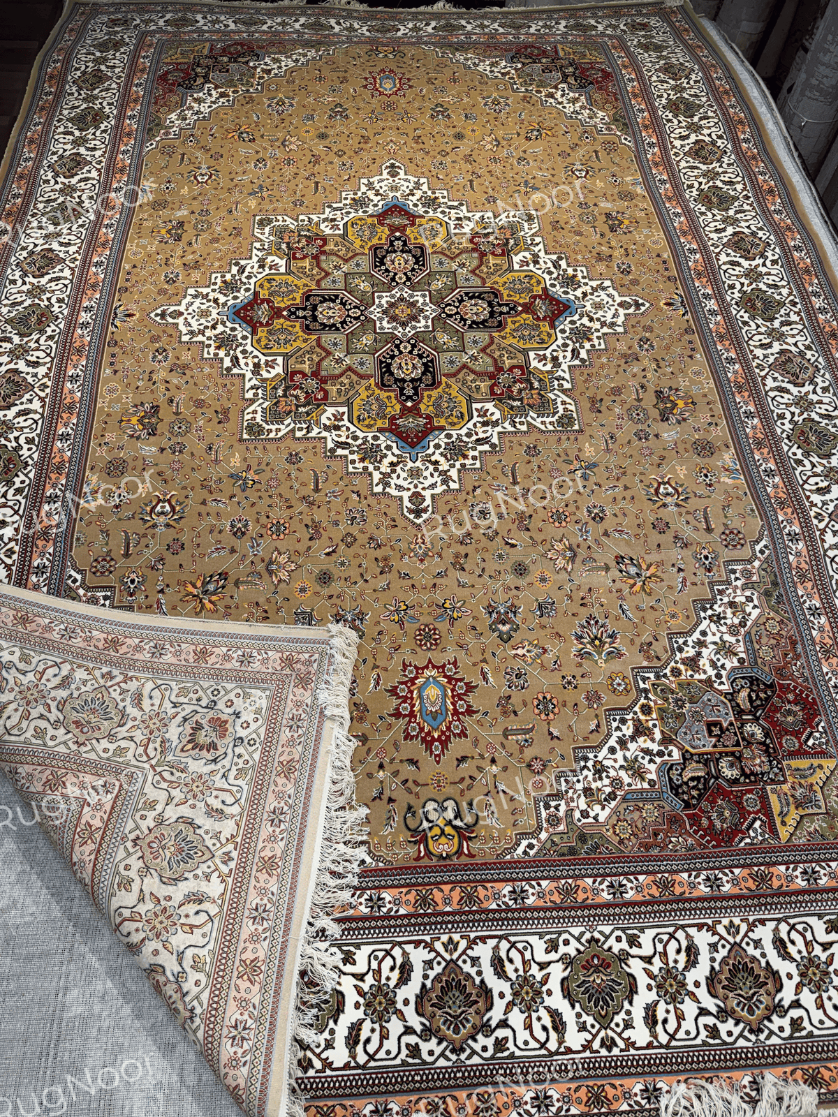 Anbara rectangular carpet with one corner folded back, showing the patterned front, pale underside, ivory fringe, and layered ornamental borders.