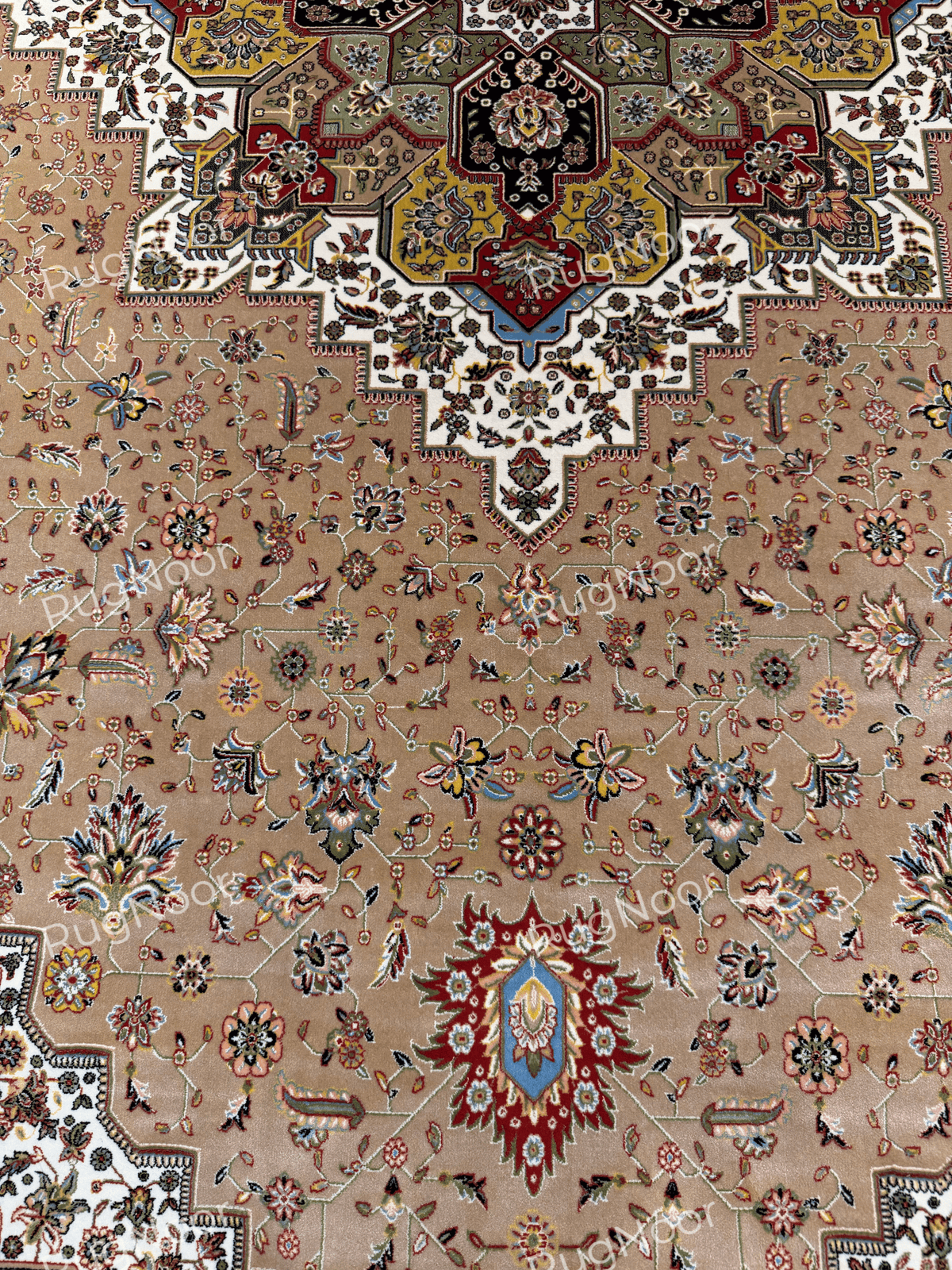 Detail of the Anbara carpet showing the lower portion of its ornate central medallion, scattered floral motifs, and a small red, blue, and ivory decorative motif on the camel-beige field.