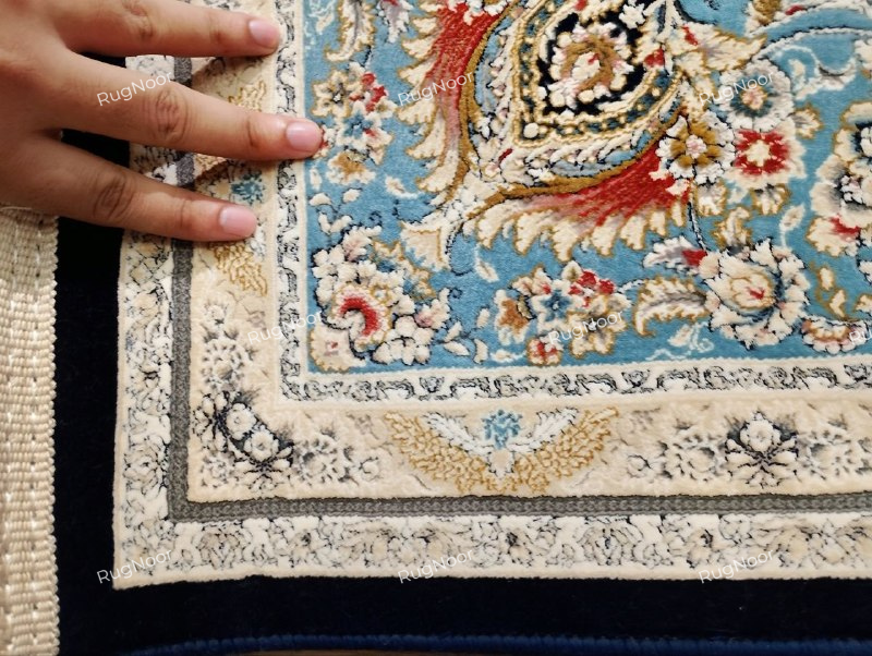 Close-up of the Lajvard rug’s layered ivory and light blue floral borders, dark edging, cream fringe, and multicoloured floral details, with a hand showing the rug’s edge.