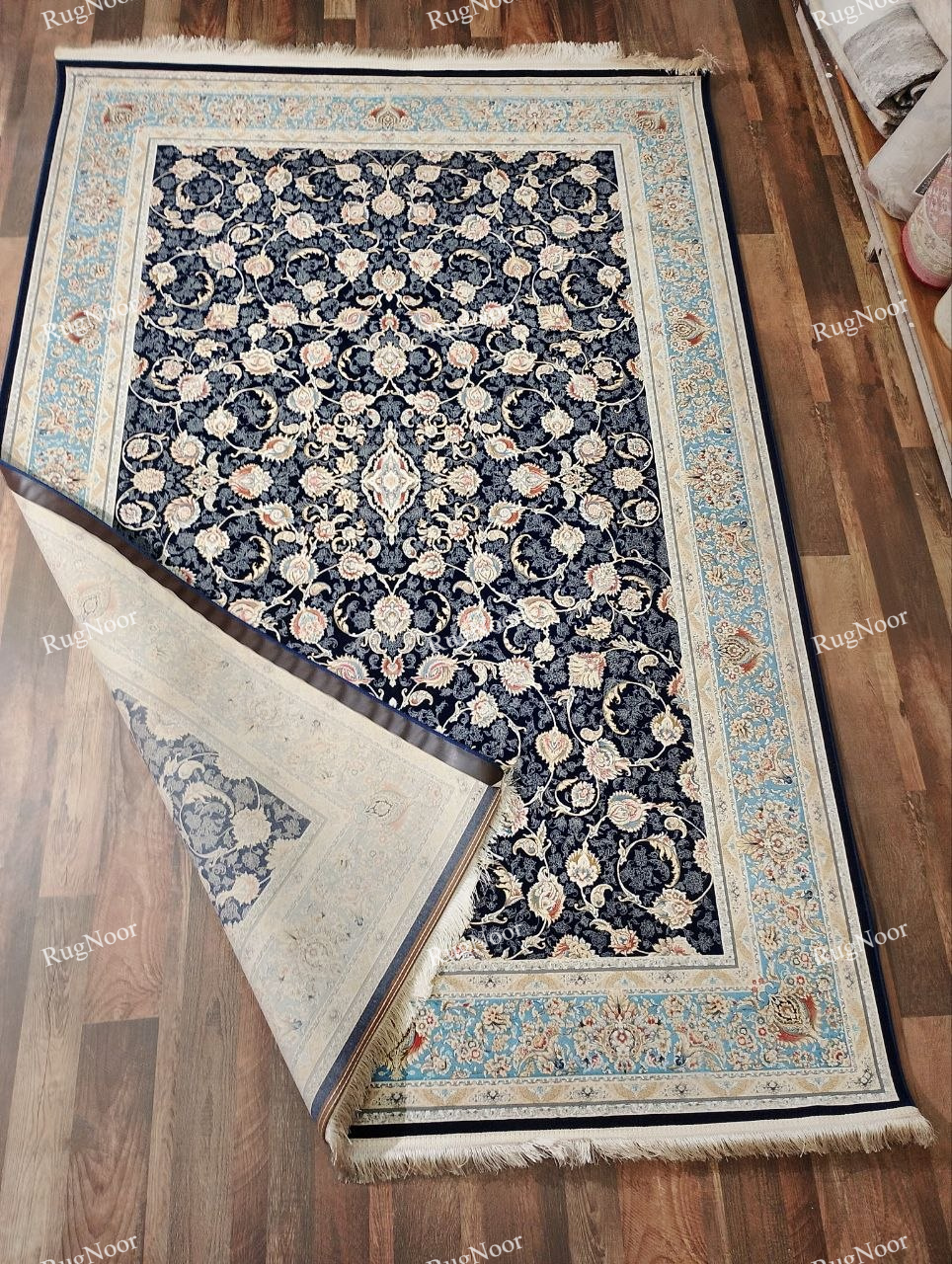 Lajvard navy blue floral rug with one corner folded back to reveal the underside, alongside the all-over floral pattern, light blue ornamental border, and cream fringe.
