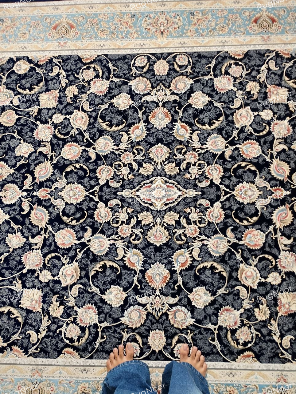 Top view of the Lajvard rug’s dense ivory, blush, and muted gold floral motifs linked by scrolling vines across a dark navy field, framed by a light blue and ivory border.