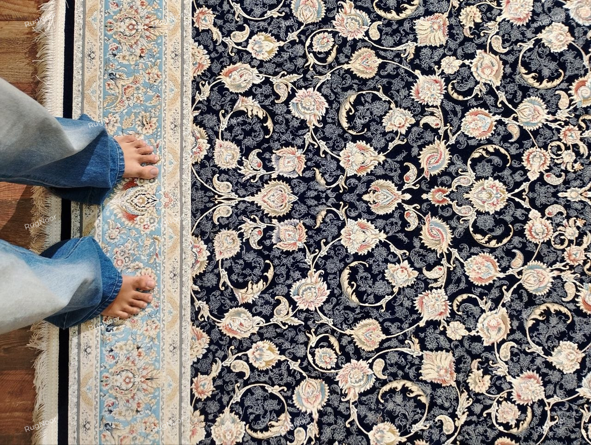 Angled view of the Lajvard rug showing its light blue and ivory side border, dark navy floral field, intricate scrolling motifs, and the border’s contrast with the wooden floor.