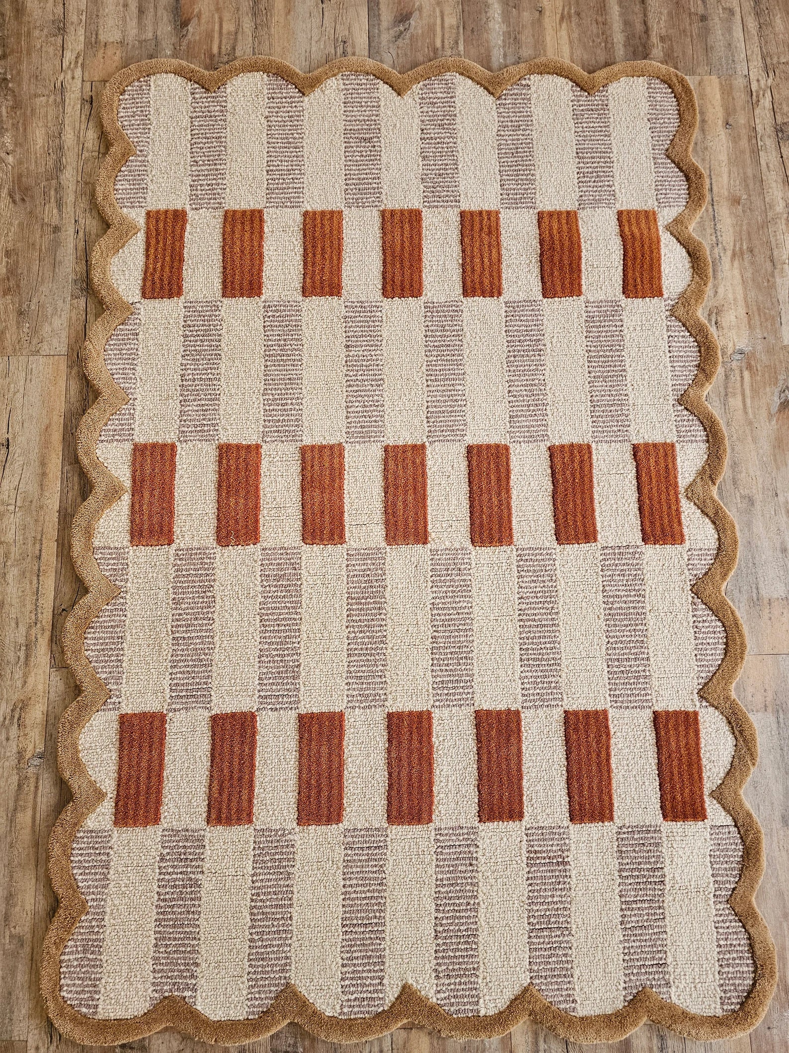 A rectangular woven rug with a scalloped jute border, featuring a checkered pattern of beige, brown, and rust-orange squares, laid flat on a rustic wooden floor.
