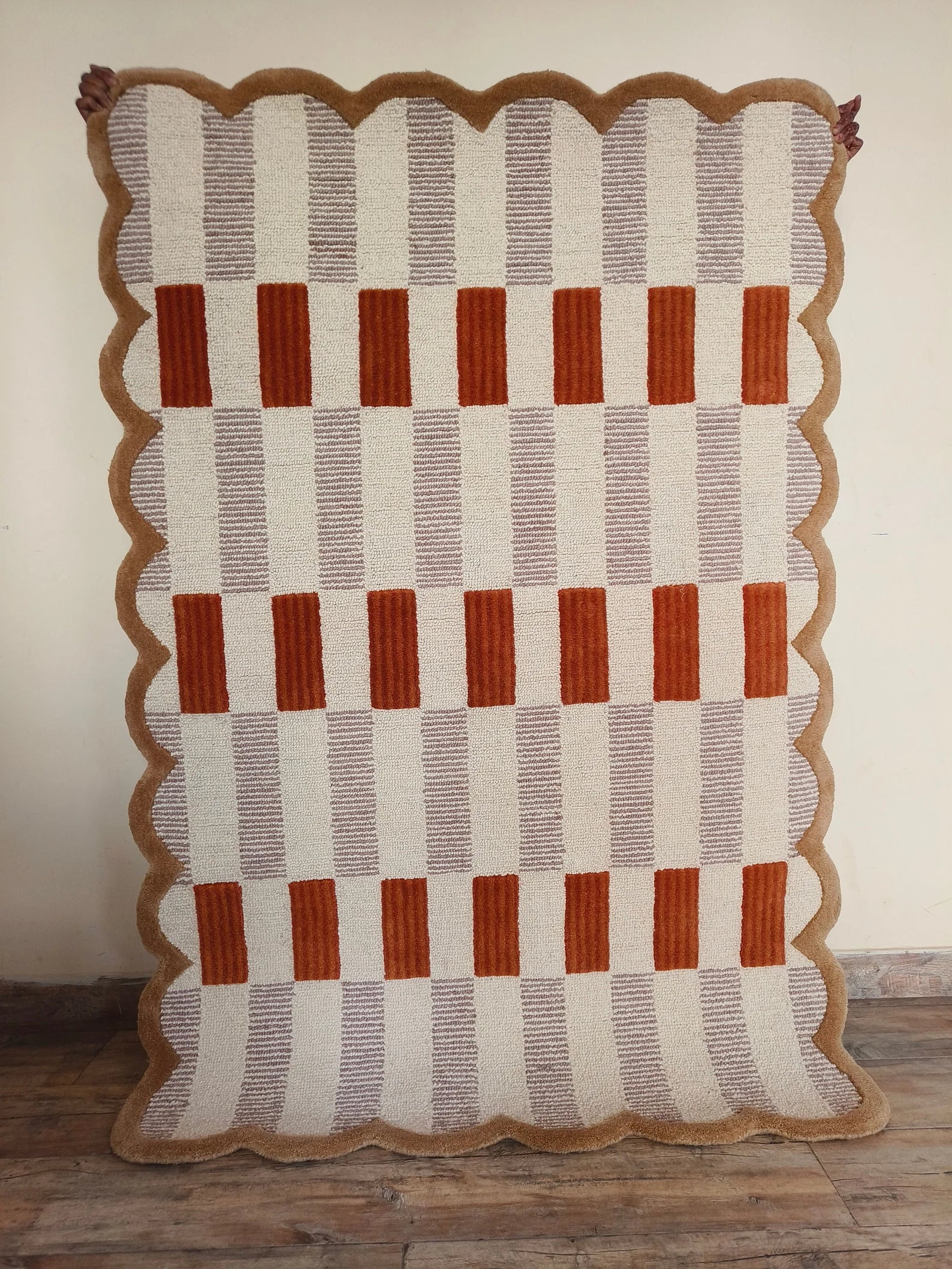 A rectangular woven textile with a scalloped brown border, featuring alternating vertical cream and taupe stripes with rust-orange rectangular blocks, displayed against a plain wall on a wooden floor.