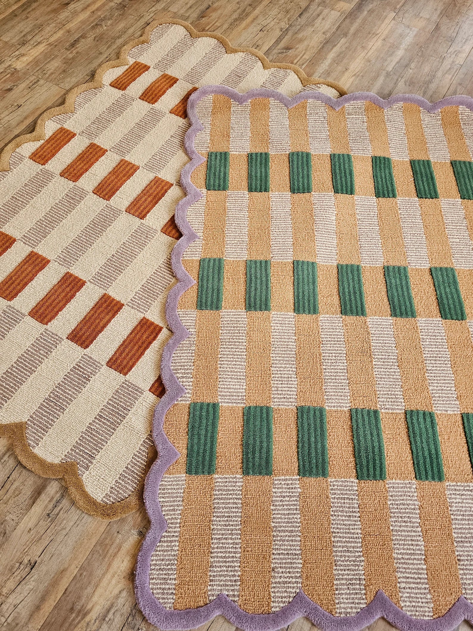 Two scalloped woven rugs laid on a wood floor. The front rug has a tan, cream, and green check pattern with a lavender border, and the rear rug has a tan, cream, and rust-orange check pattern with a tan border.