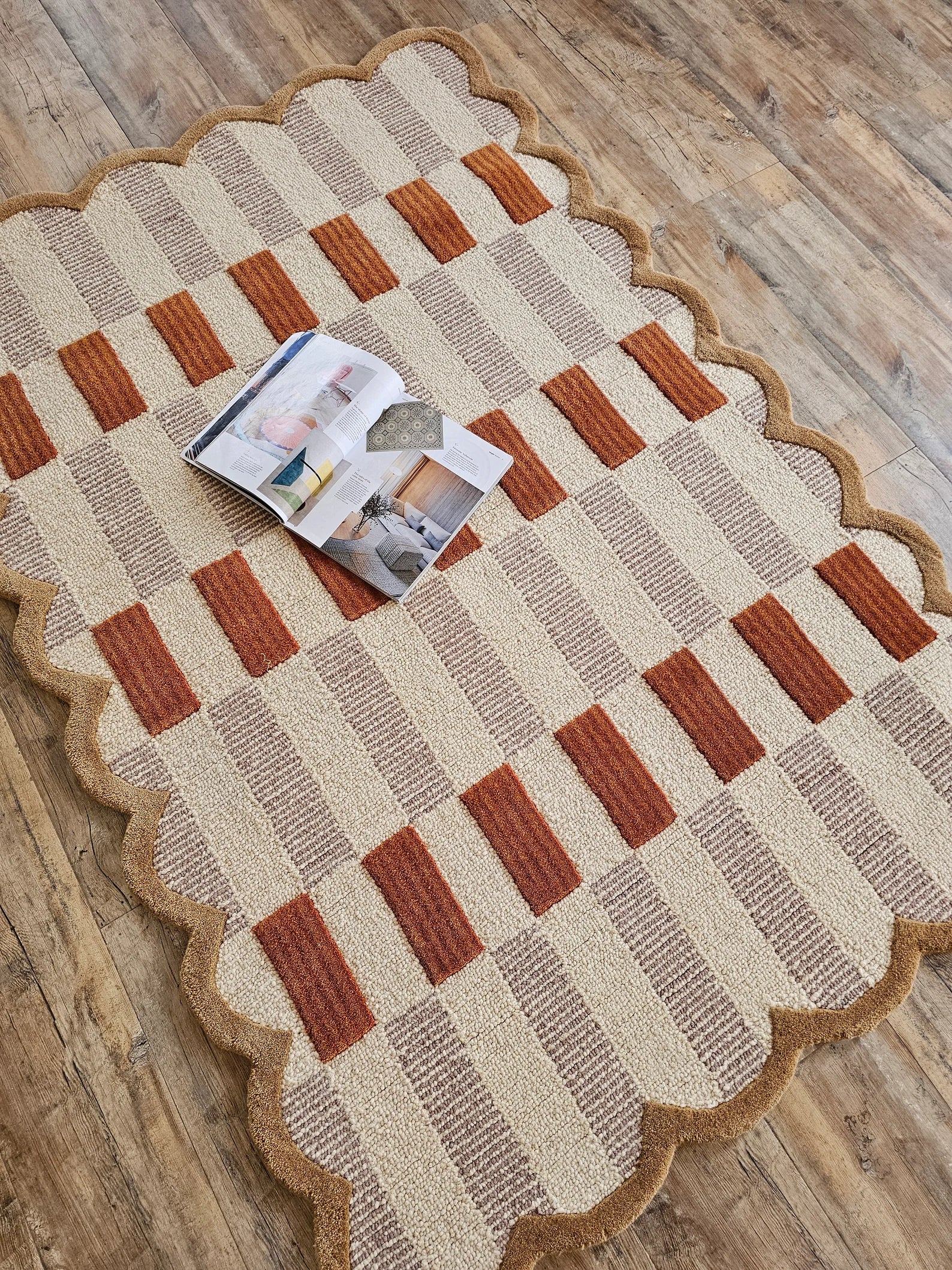 A rectangular area rug with a scalloped edge, featuring a textured checkerboard pattern in beige, taupe, and rust orange, laid out on a rustic wooden floor with an open magazine resting on it.