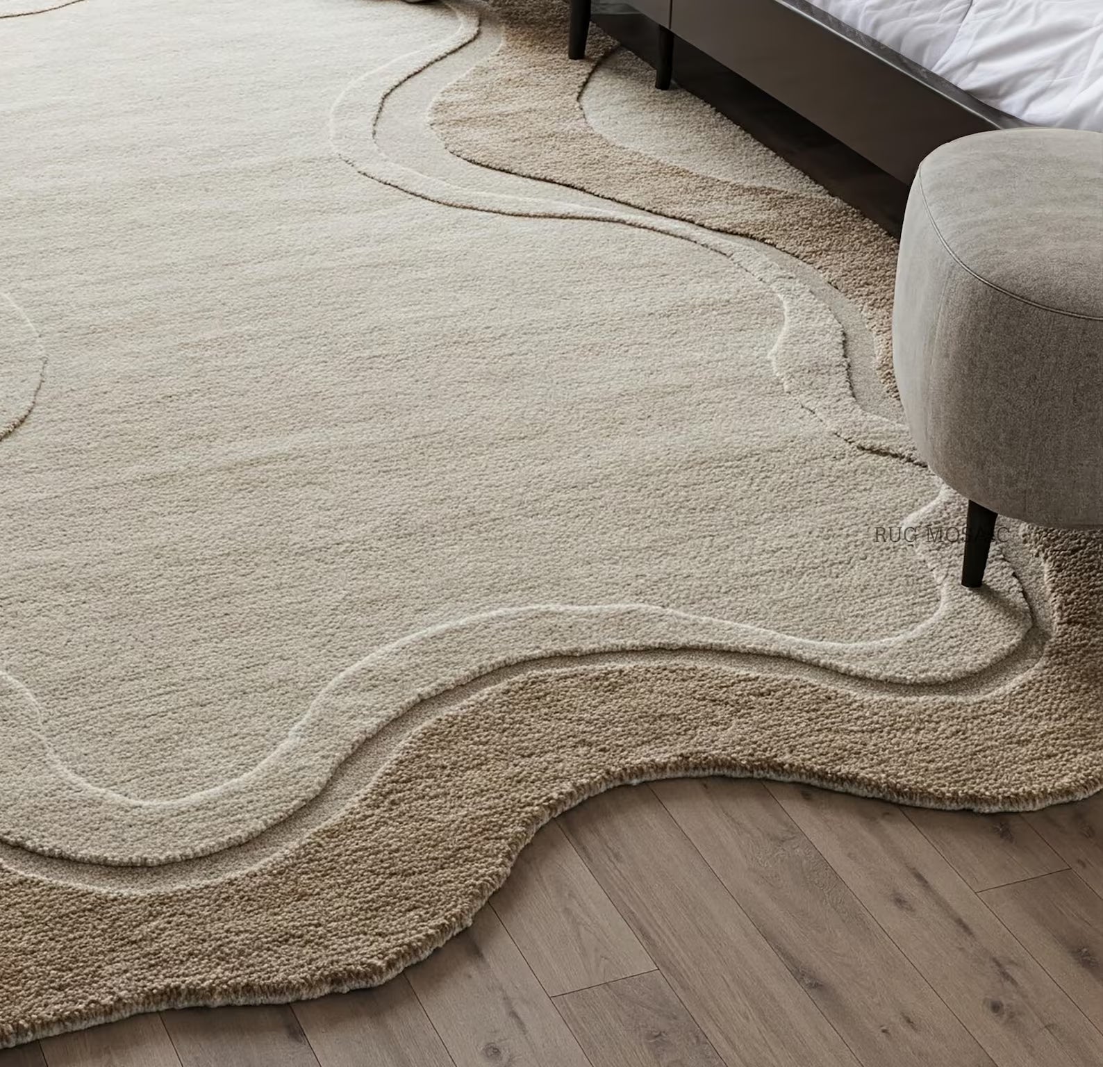 A large abstract area rug in warm beige and cream tones with flowing wavy lines, shown on a light wood floor in a bedroom setting with part of a bed and a round upholstered ottoman visible.