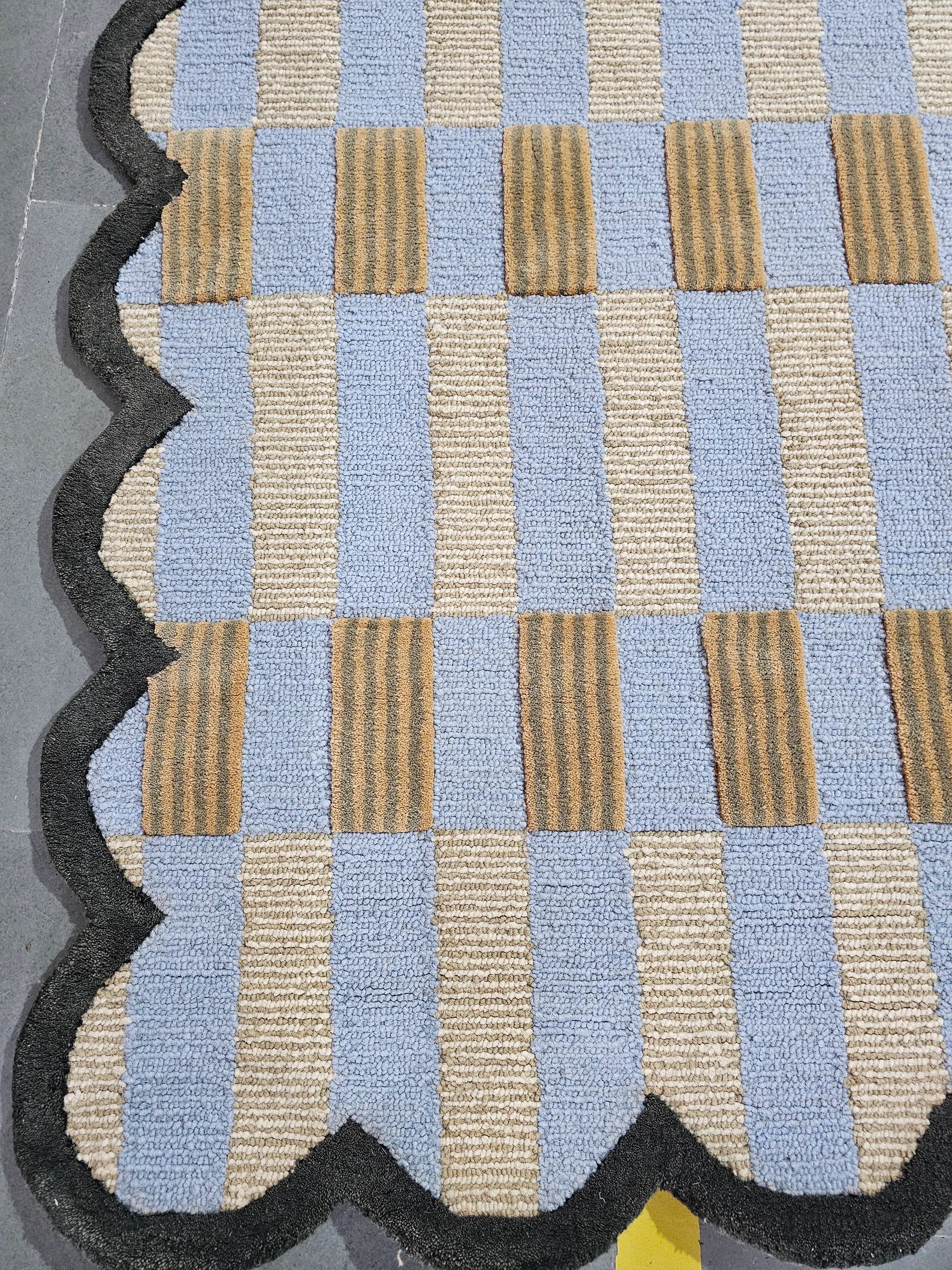 A close-up of a textured area rug with a woven checkerboard pattern of light blue, beige, and brown striped squares, finished with a scalloped dark gray border.