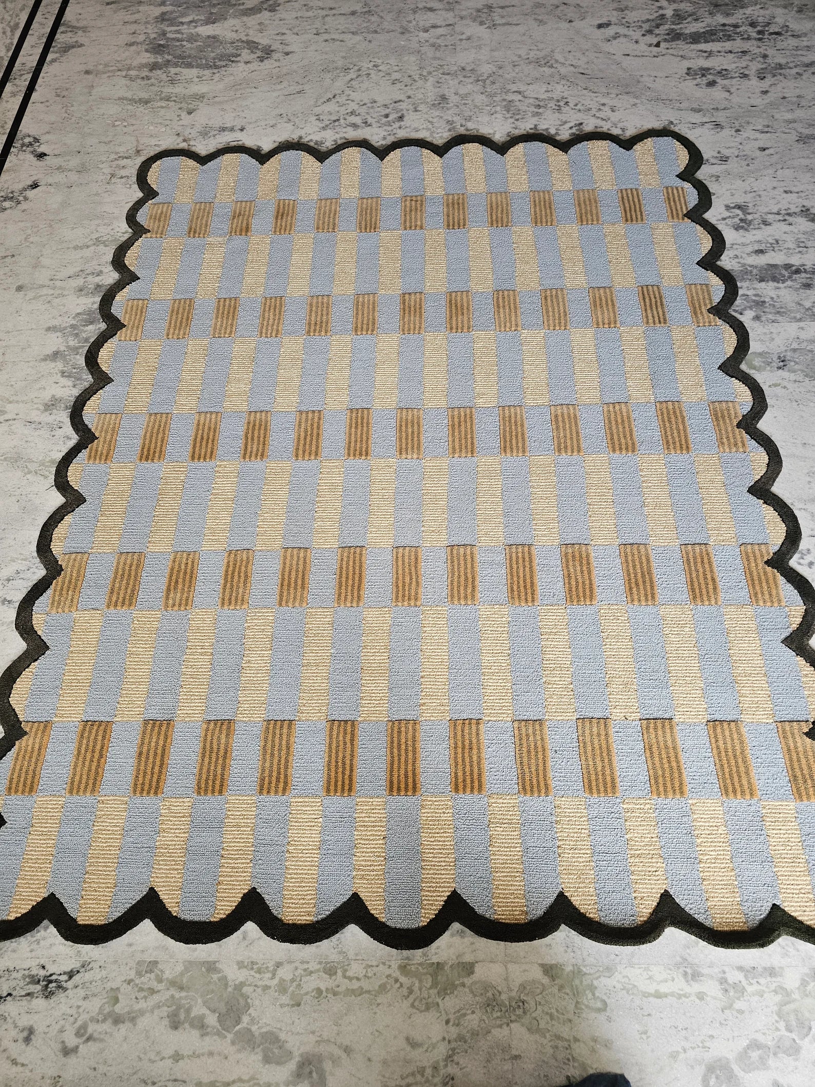 A rectangular woven rug with a scalloped black edge, featuring a checkered pattern of light blue and beige squares with vertical tan stripes, laid flat on a gray concrete floor.
