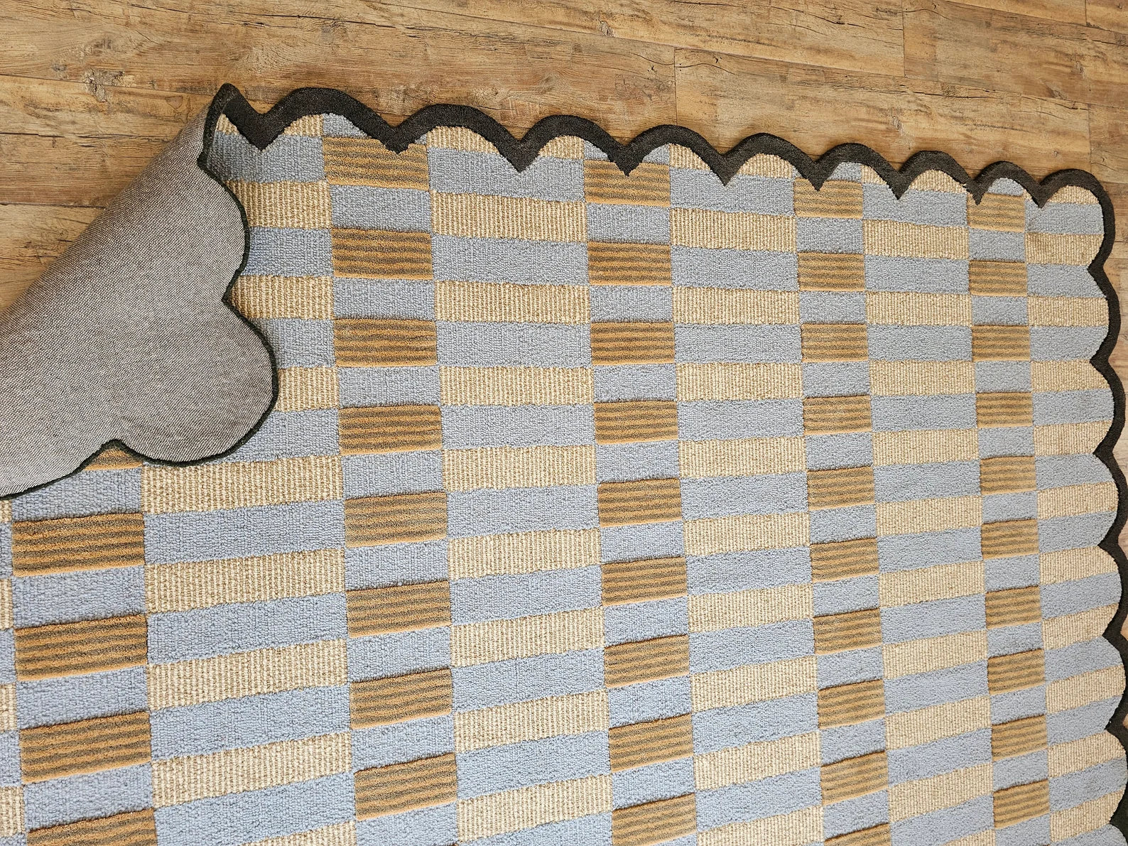 A checkered area rug with a scalloped black edge, shown partially unrolled on a light wood floor. The rug features alternating squares of light blue-gray, natural tan, and beige, with a gray backing visible on the underside.
