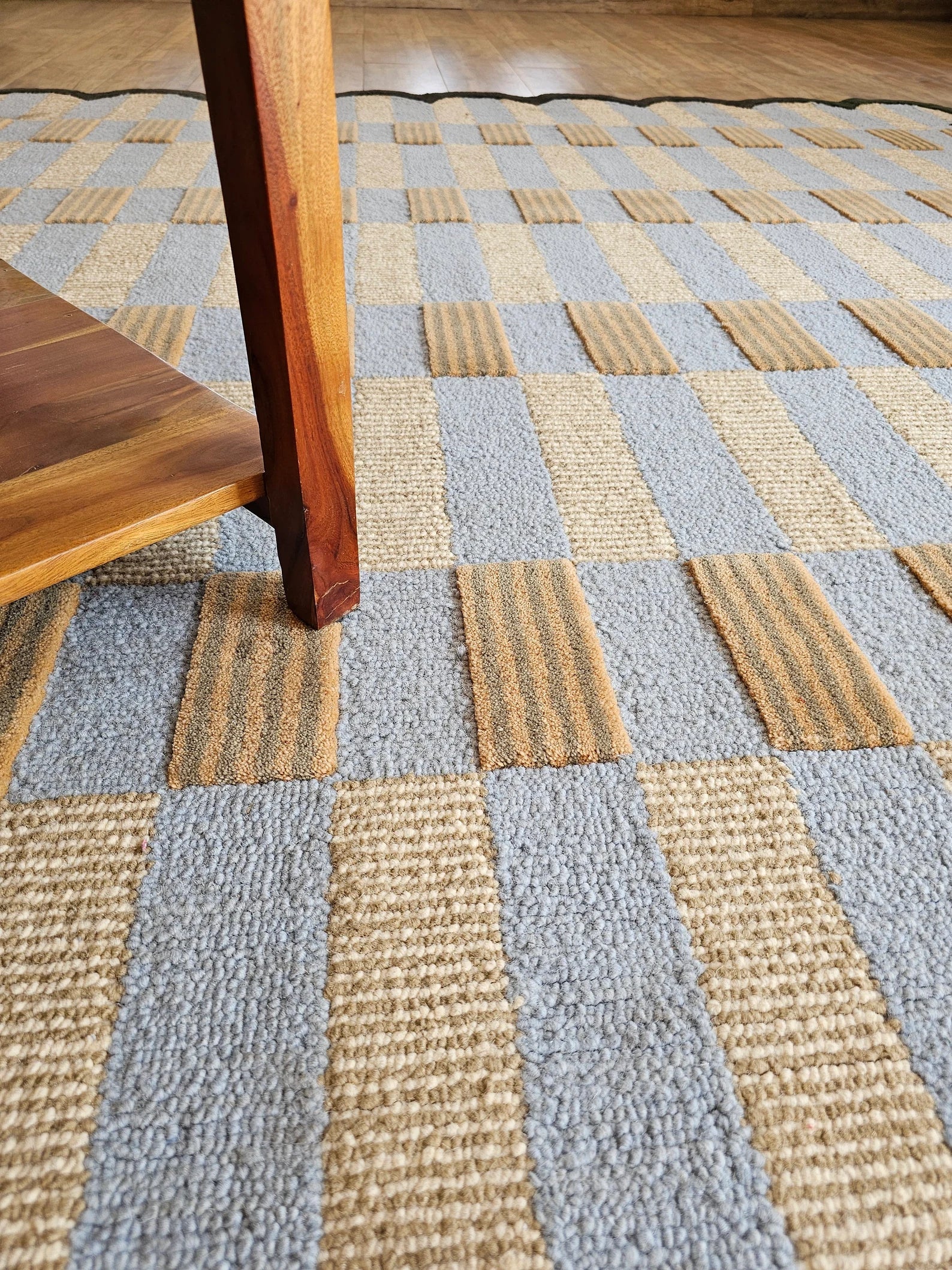 A close-up of a woven area rug with a checkered pattern in light blue-gray and natural tan, with a wooden table leg visible in the foreground.