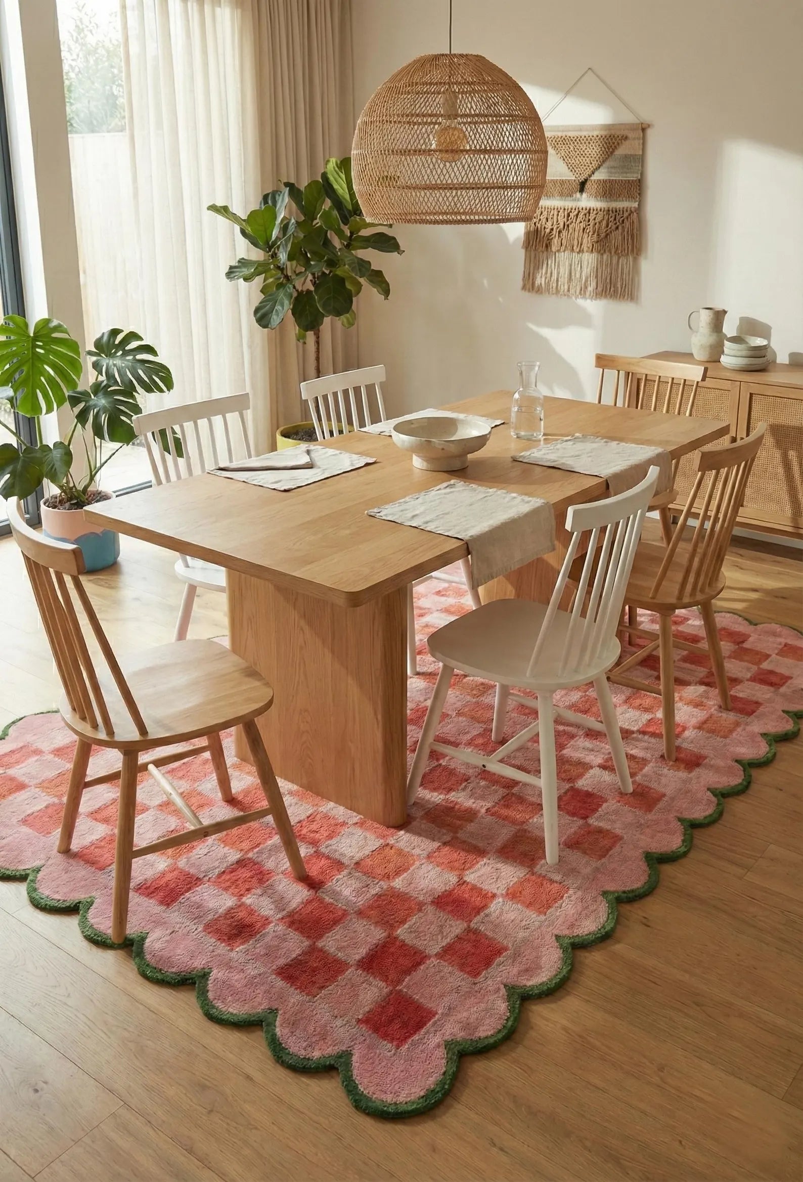 A styled dining room scene featuring a light wood dining table and chairs on a pink and red checkered scalloped area rug with a green border, set in a warm, sunlit room with plants and a wicker pendant light.