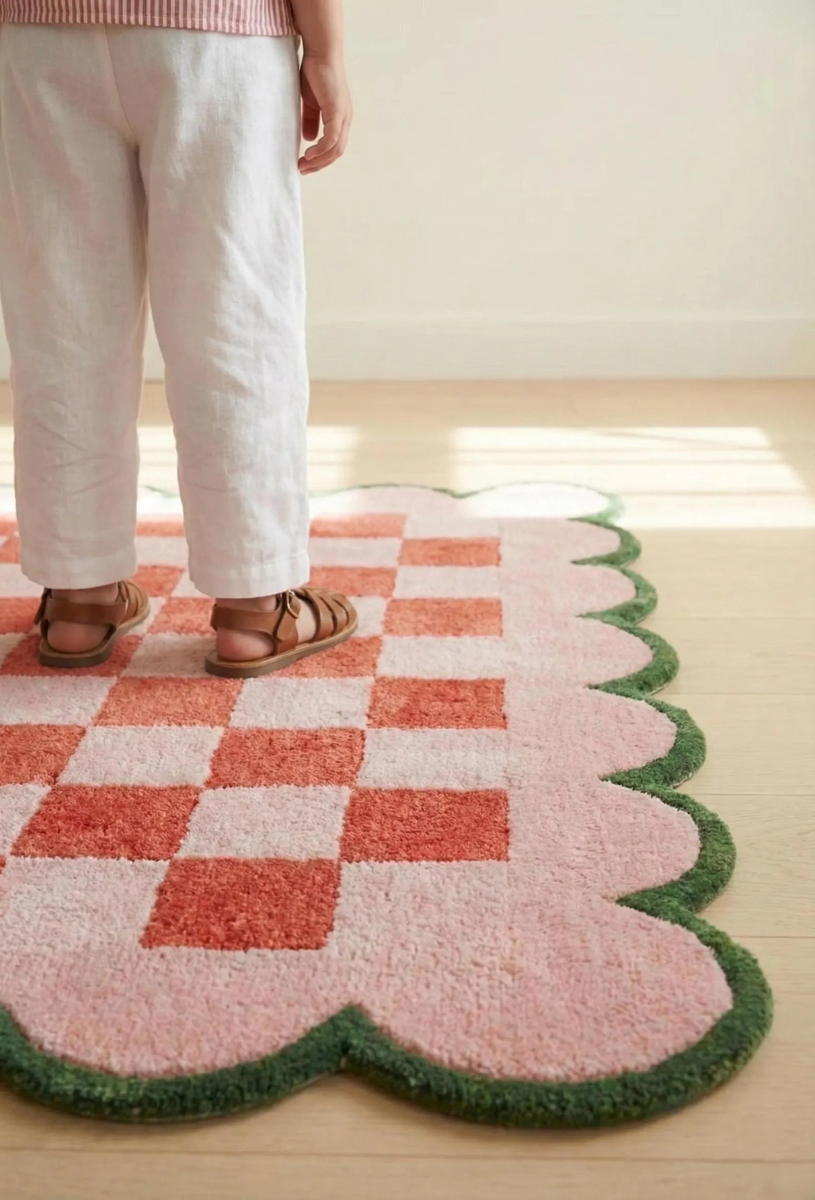 A child wearing white linen pants and brown sandals stands on a pink and white checkered rug with a scalloped green border on a light wood floor.