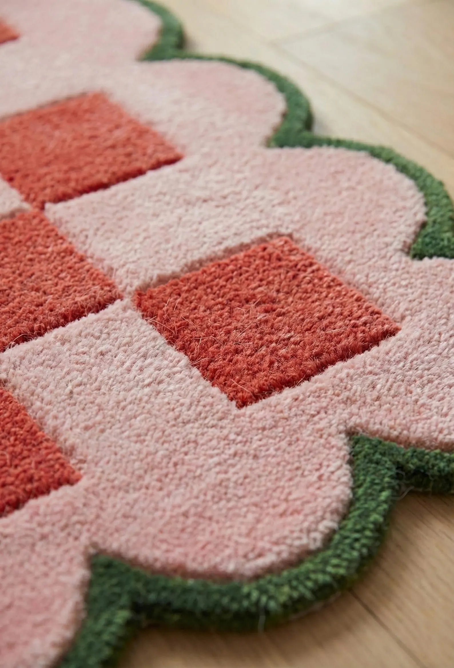 A close-up of a textured area rug with a pink and coral-red geometric pattern and a green scalloped border, shown on a light wood floor.