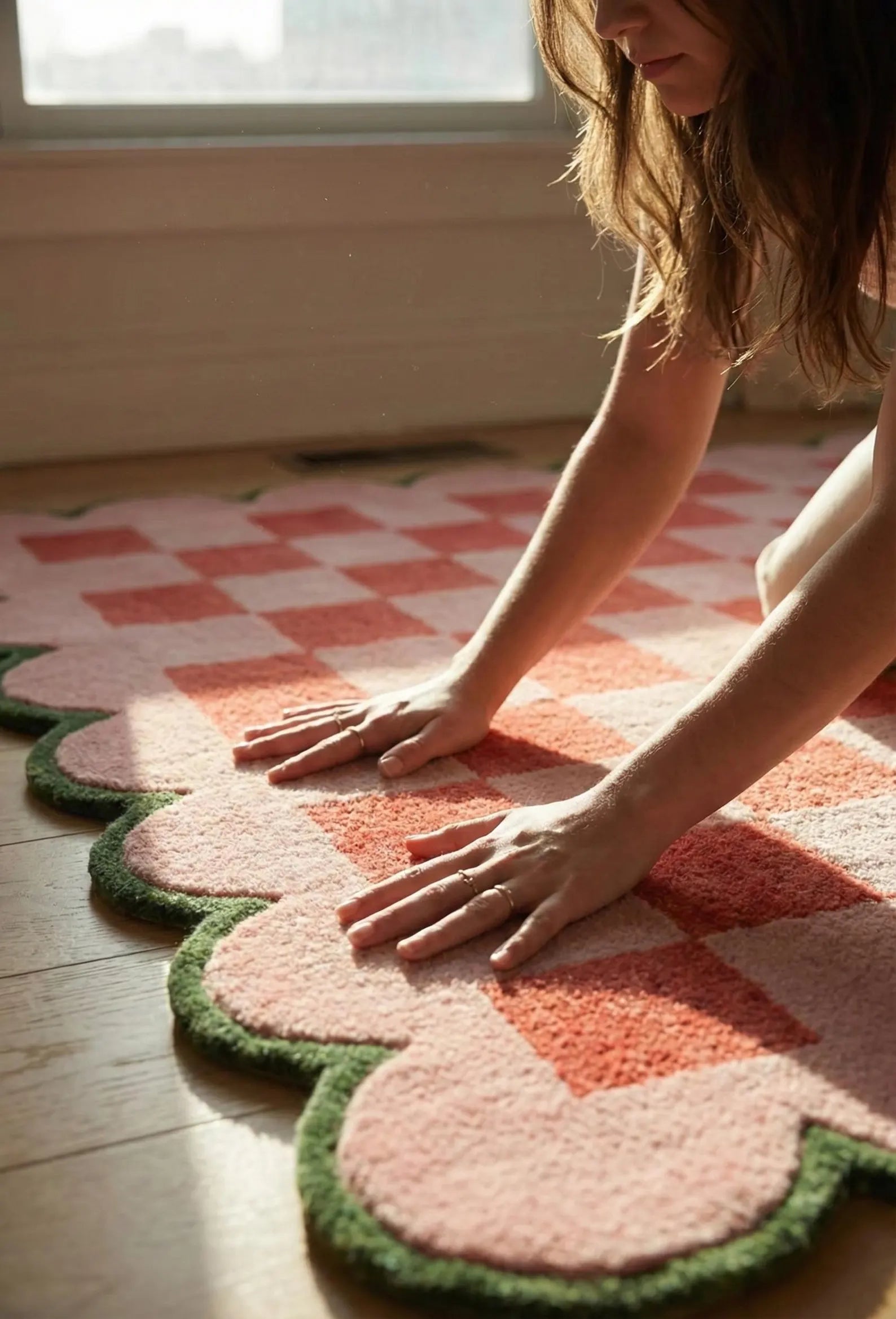 A woman kneels on a pink and coral checkered rug with a green scalloped border on a wooden floor, with warm sunlight streaming in from a window.