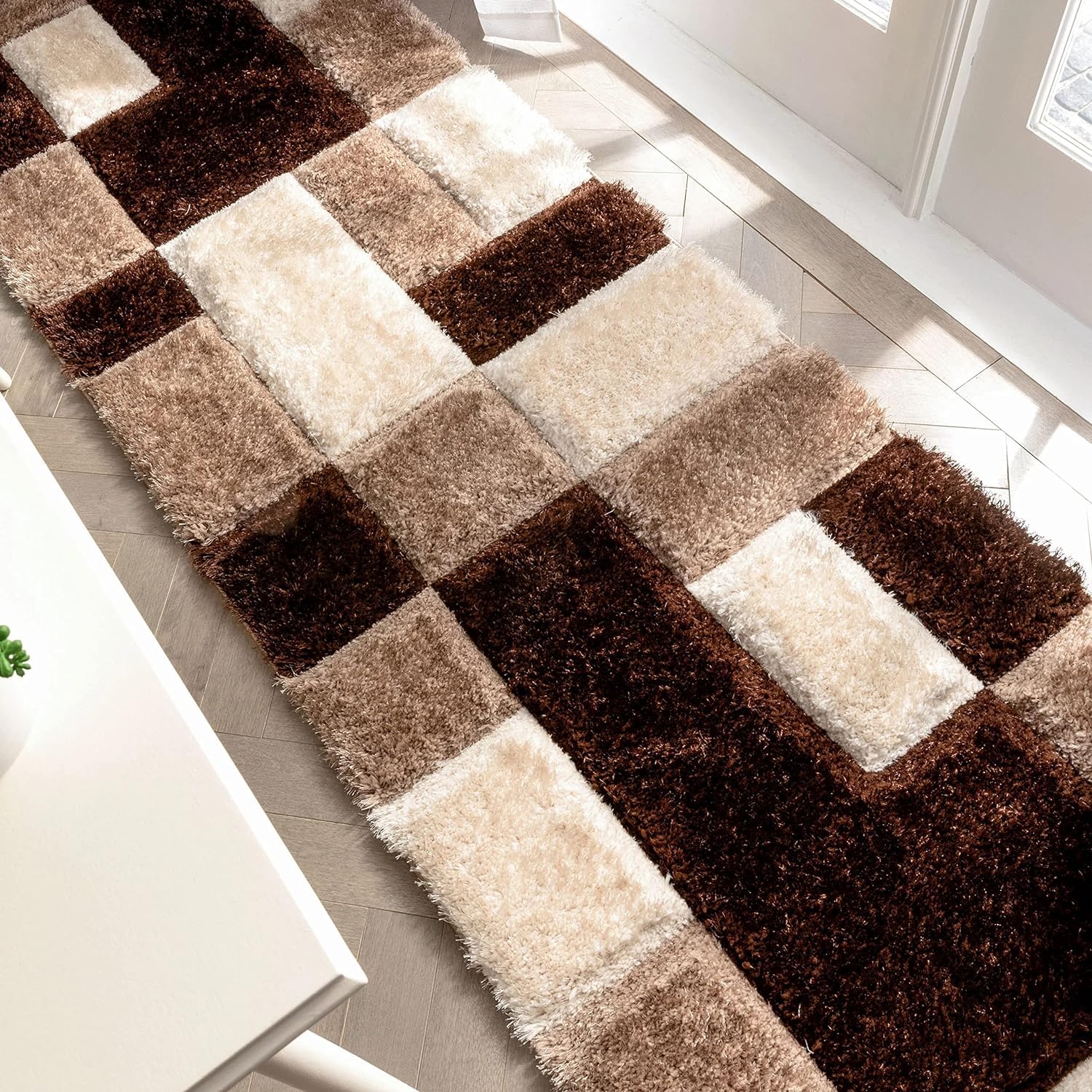 A rectangular shag area rug with a geometric patchwork pattern in dark brown, medium brown, beige, and cream, shown on a light-colored tiled floor in a bright room.