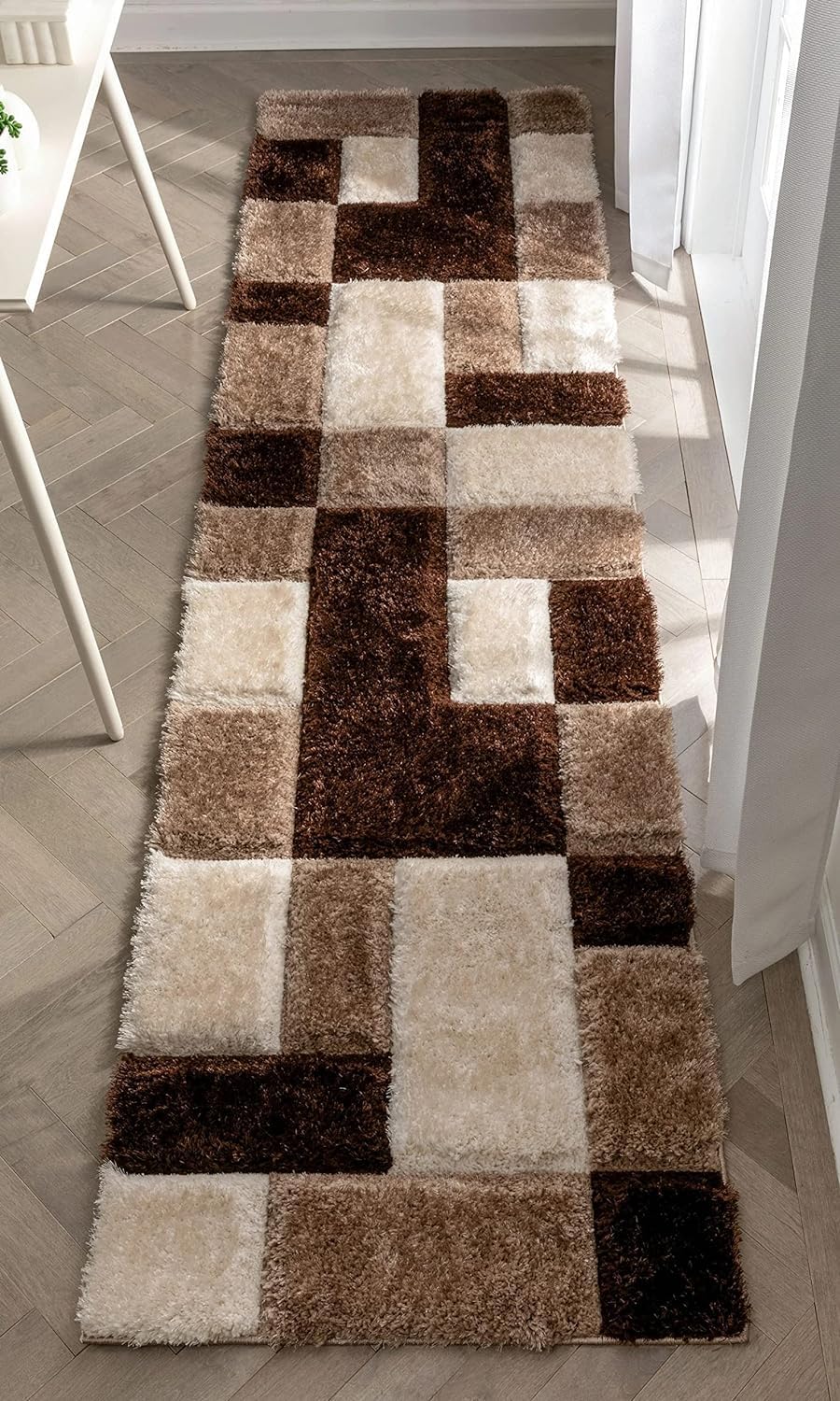 A long rectangular shag runner rug with a geometric patchwork pattern in shades of brown, beige, and cream, shown on a light wood floor in a bright room.
