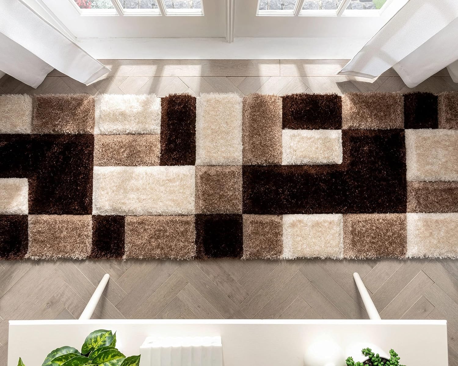 A rectangular shag area rug with a geometric block pattern in shades of brown, beige, and cream, shown on a light wood herringbone floor in a bright room.