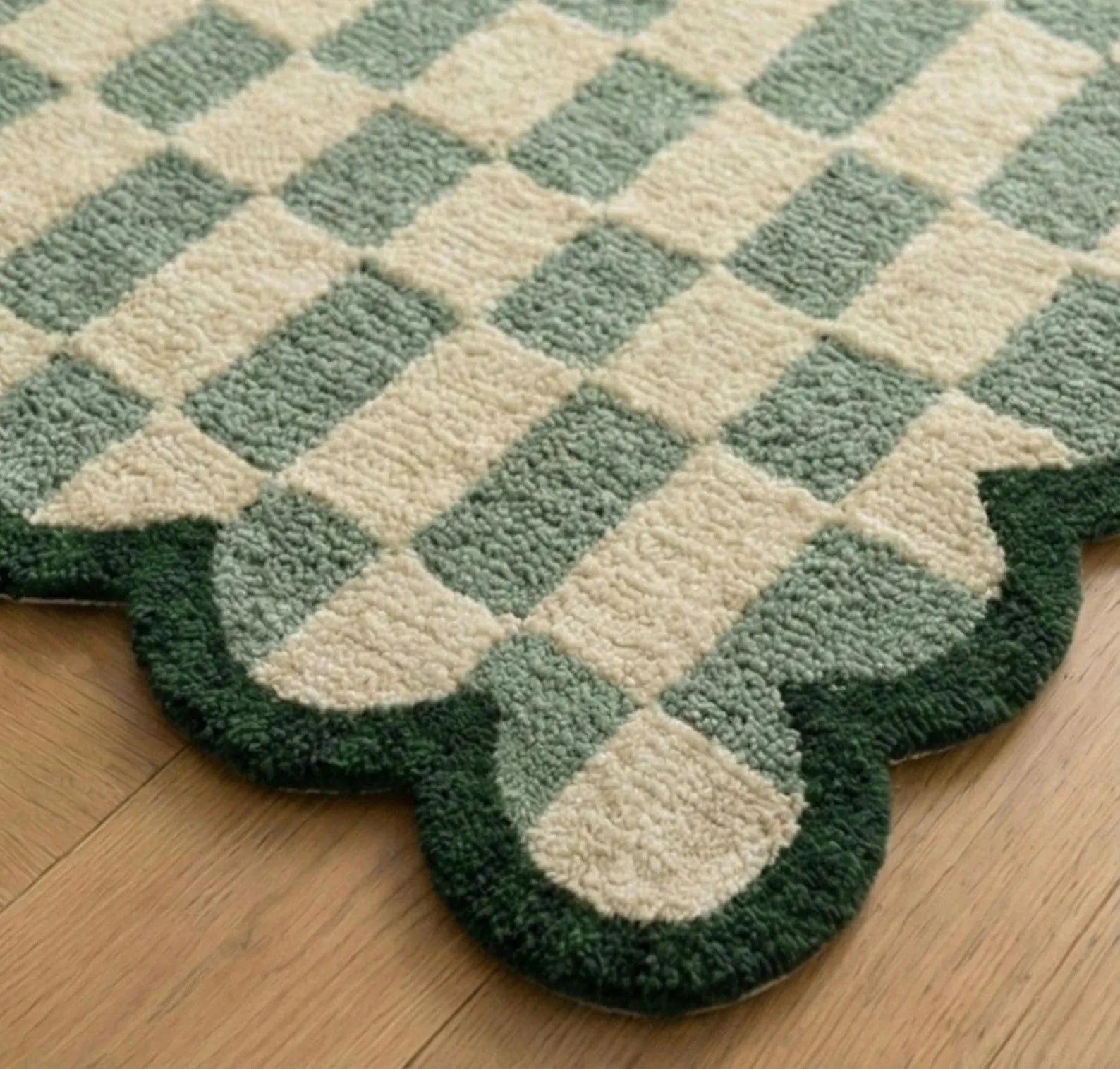 A close-up of a textured area rug with a green and cream checkerboard pattern and a scalloped dark green border, shown on a light wood floor.