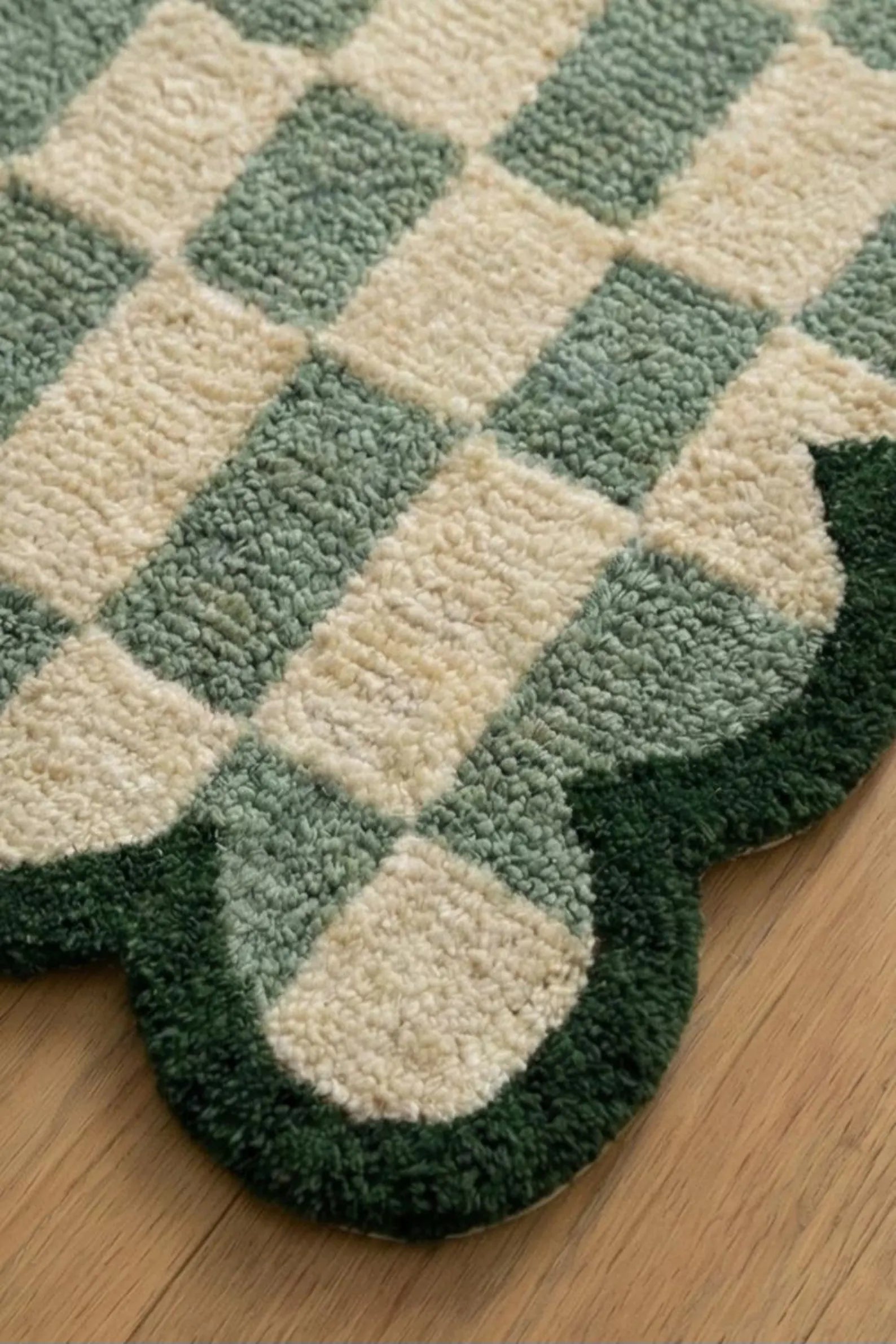 A close-up of a textured area rug with a green and cream checkerboard pattern and a scalloped edge, shown on a light wood floor.