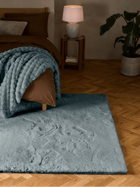 A plush bluish gray area rug in a styled bedroom setting with a wooden herringbone floor, a bed with a chunky knit blue throw blanket, a bedside table with a lamp, and a potted plant.