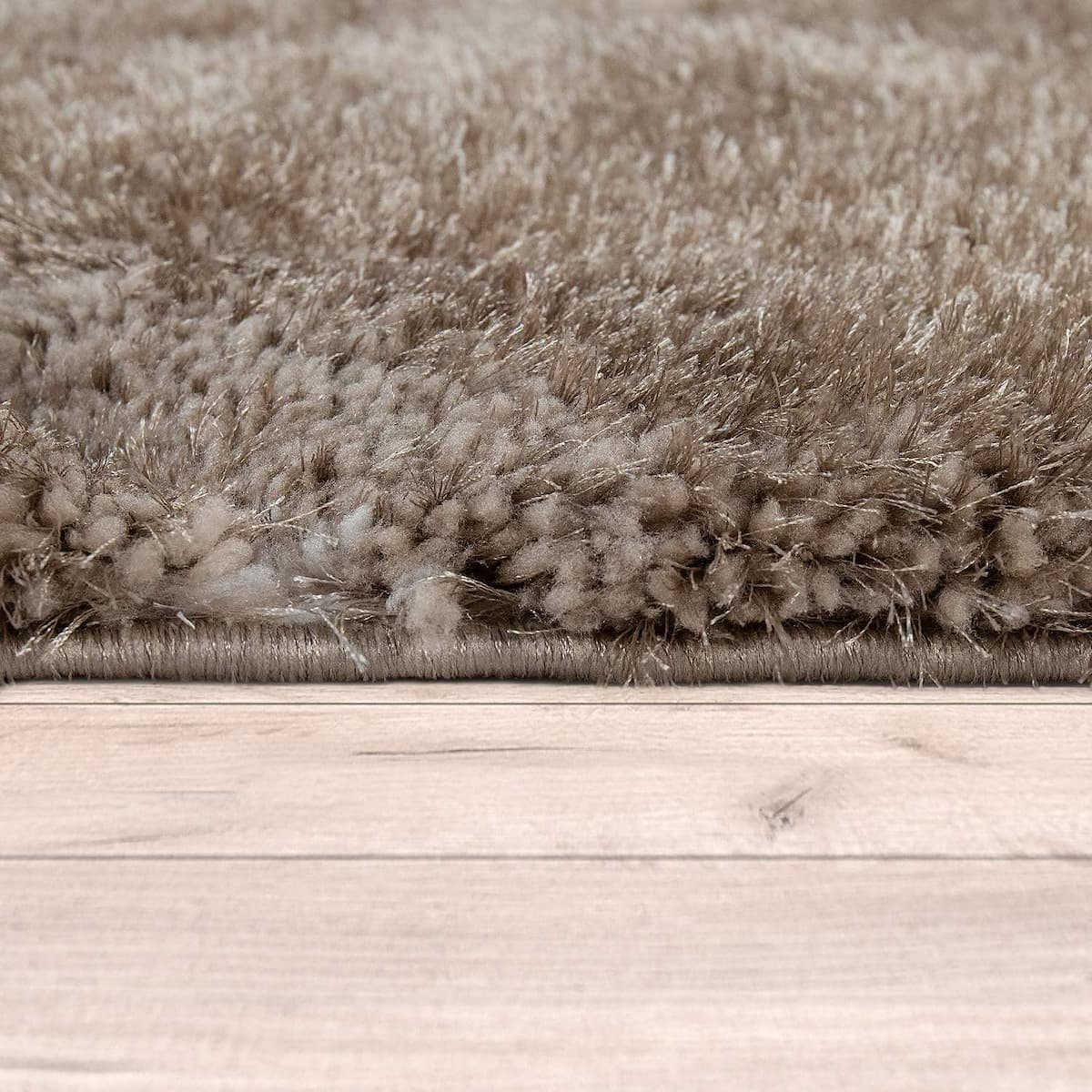 Close-up of a shaggy taupe area rug with a thick pile, shown folded over to reveal the edge and texture, resting on a light wood floor.