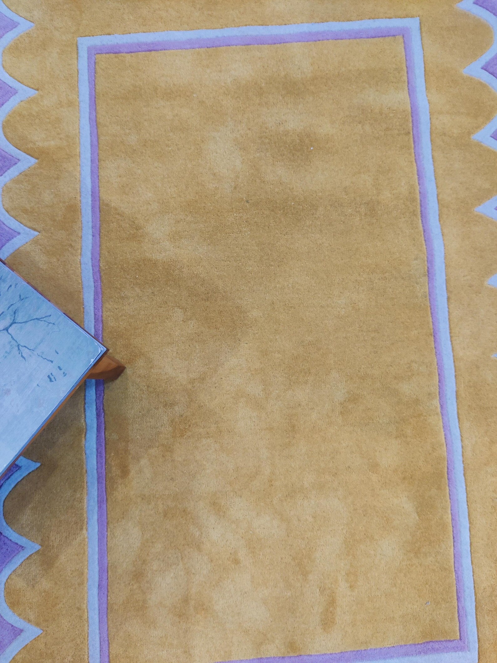 A close-up of a tan or camel-colored rug with a lavender and white border, shown from a top-down angle with a small portion of a blue patterned textile visible at the left edge.
