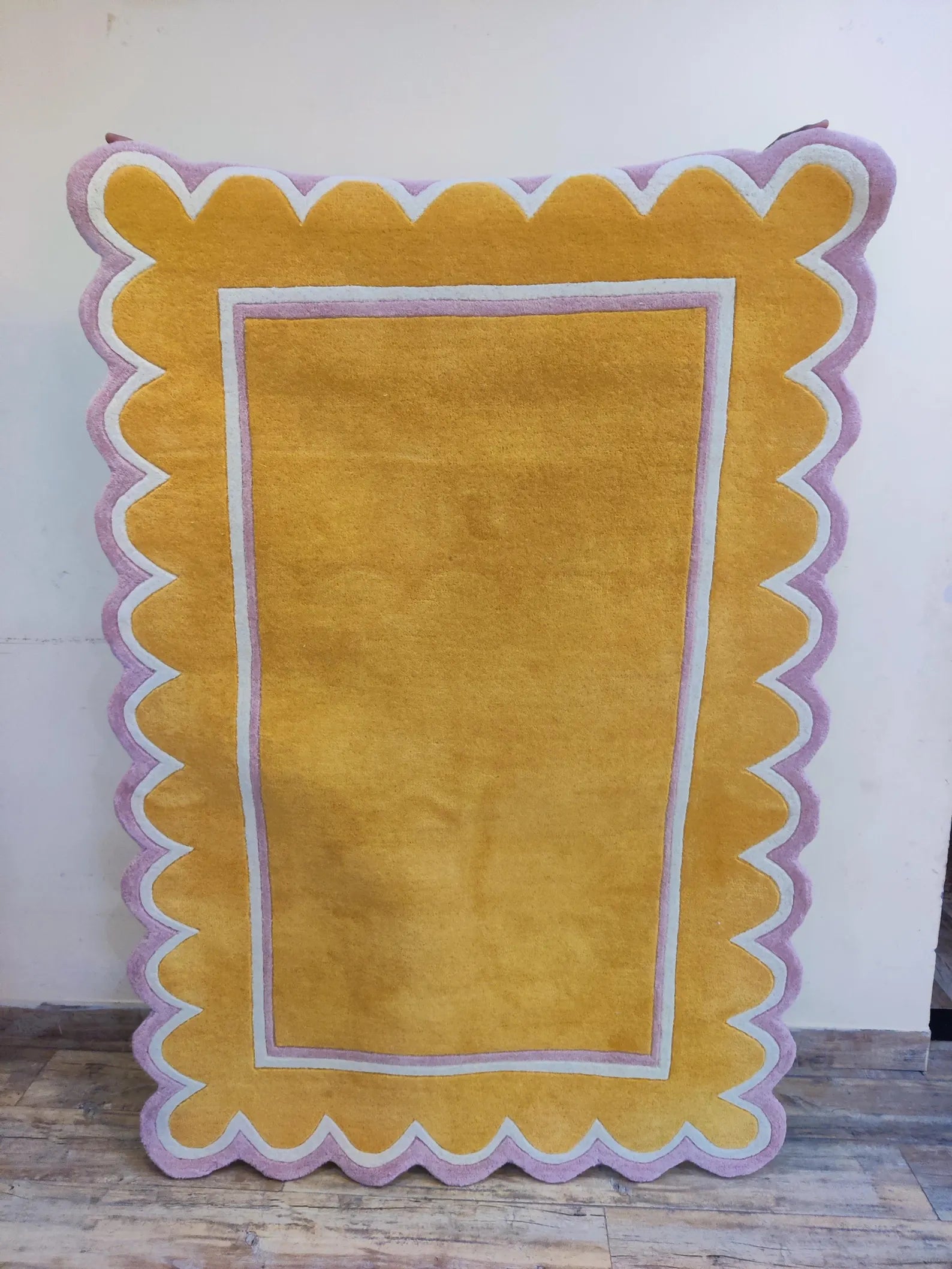 A rectangular yellow rug with a scalloped lavender and white border, displayed against a plain wall on a wooden floor.
