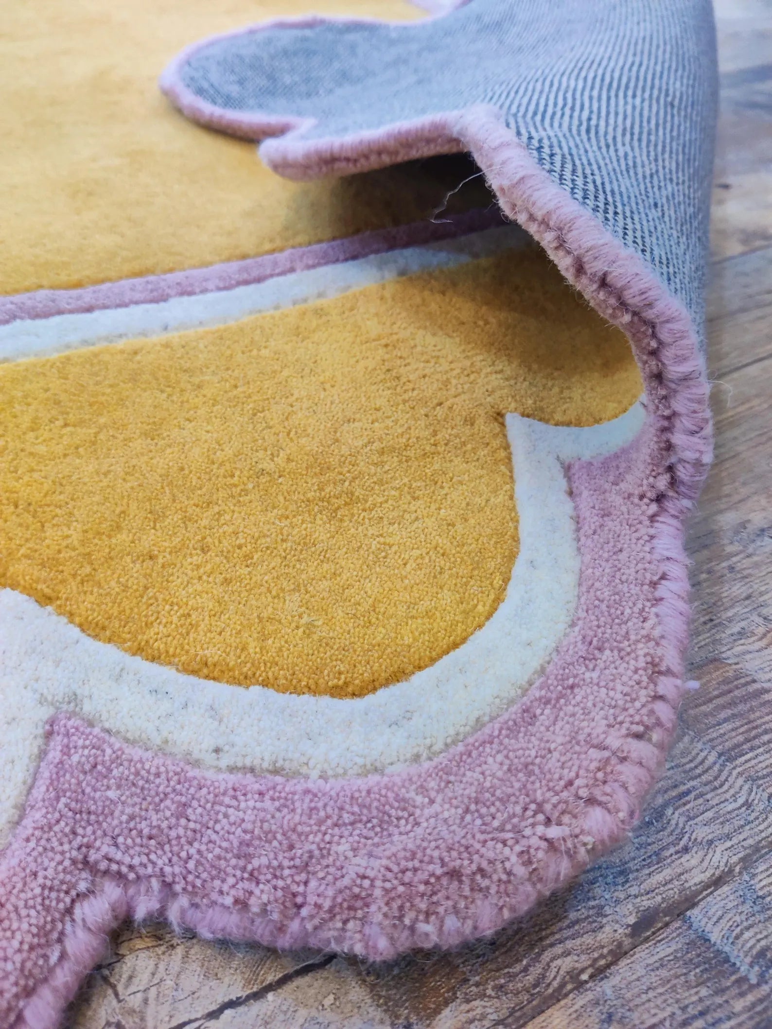 A close-up of a plush, multi-colored area rug with a raised texture, showing a yellow and dusty pink design with a light blue underside, laid on a rustic wooden floor.