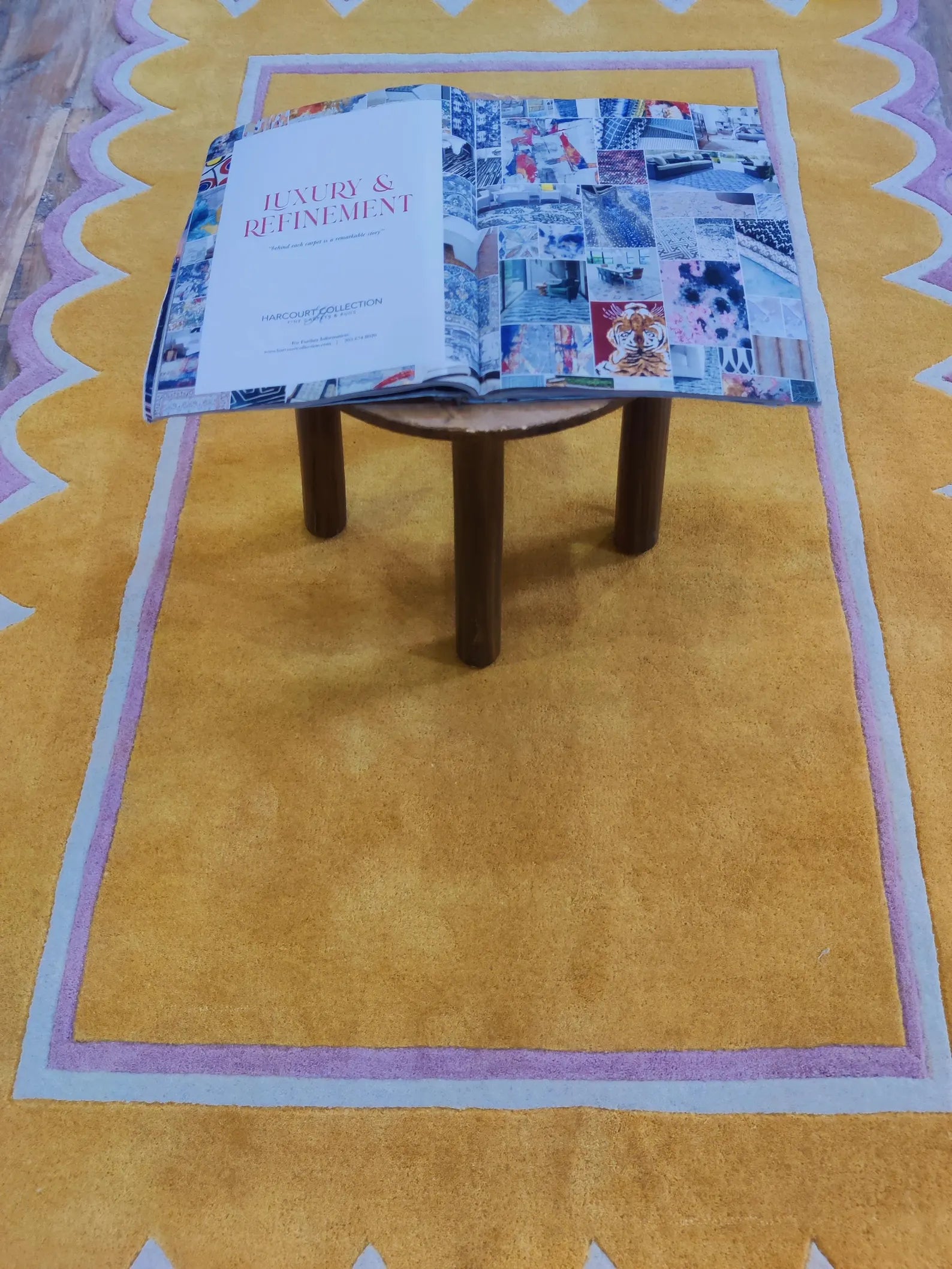 An open book titled 'LUXURY & RETIREMENT' rests on a small round wooden stool placed over a mustard yellow area rug with a pink and white wavy border.