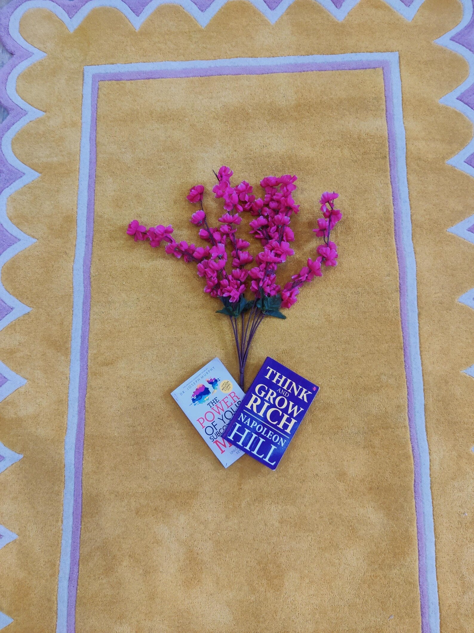 A bouquet of bright pink artificial flowers with green stems and two small cards, arranged on a tan textured rug with a purple and white border.