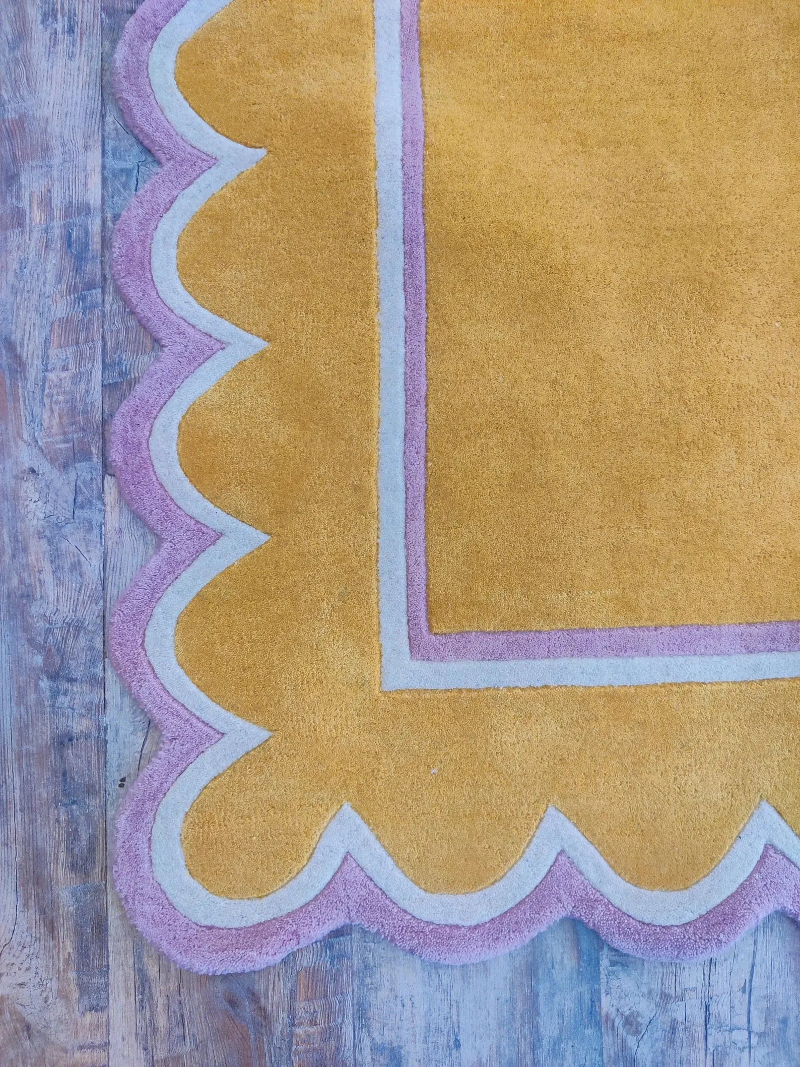A close-up of a mustard yellow rug with a scalloped border in light pink and white, laid on a rustic blue-gray wooden floor.