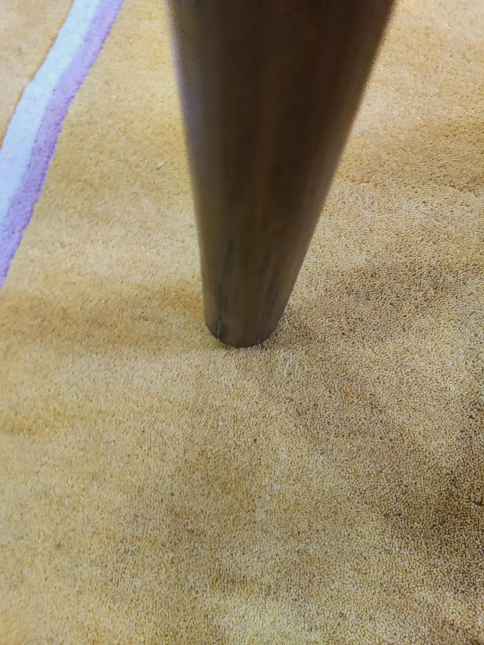 Close-up of a dark brown wooden table leg pressed into a beige textured rug with a purple stripe visible along the left edge.