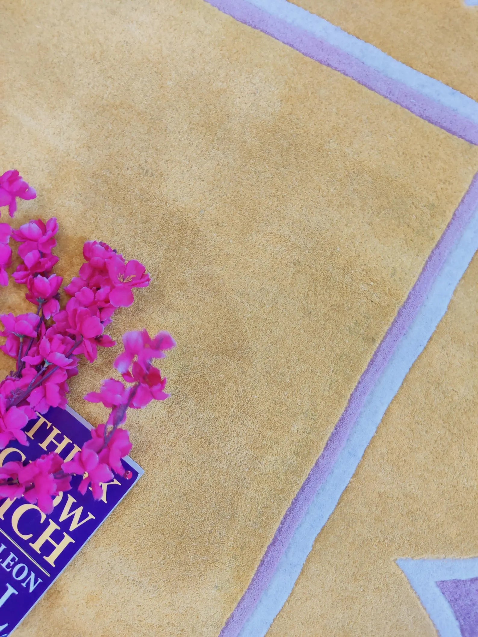 A close-up of a tan suede or suede-like fabric surface with a painted geometric border in pale lavender and light blue. Bright pink flowers and part of a purple book are placed on the lower left as styling props.